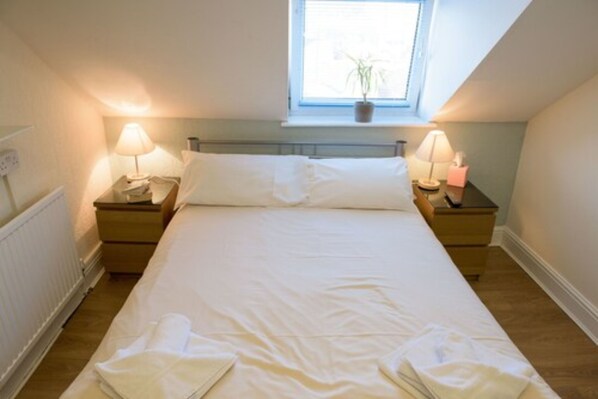 Cama arrumada em quarto com janela acima, duas mesas de cabeceira com abajures e um radiador à esquerda.