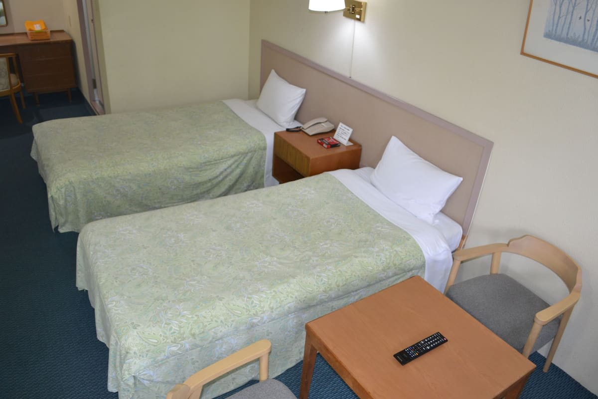 A hotel room with two single beds, a small wooden table, a chair, a nightstand with a phone and a book, and a wall-mounted lamp.