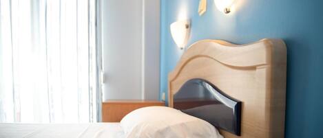 A neatly made bed with a wooden headboard and white bedding in a room with blue walls and a window with sheer curtains.