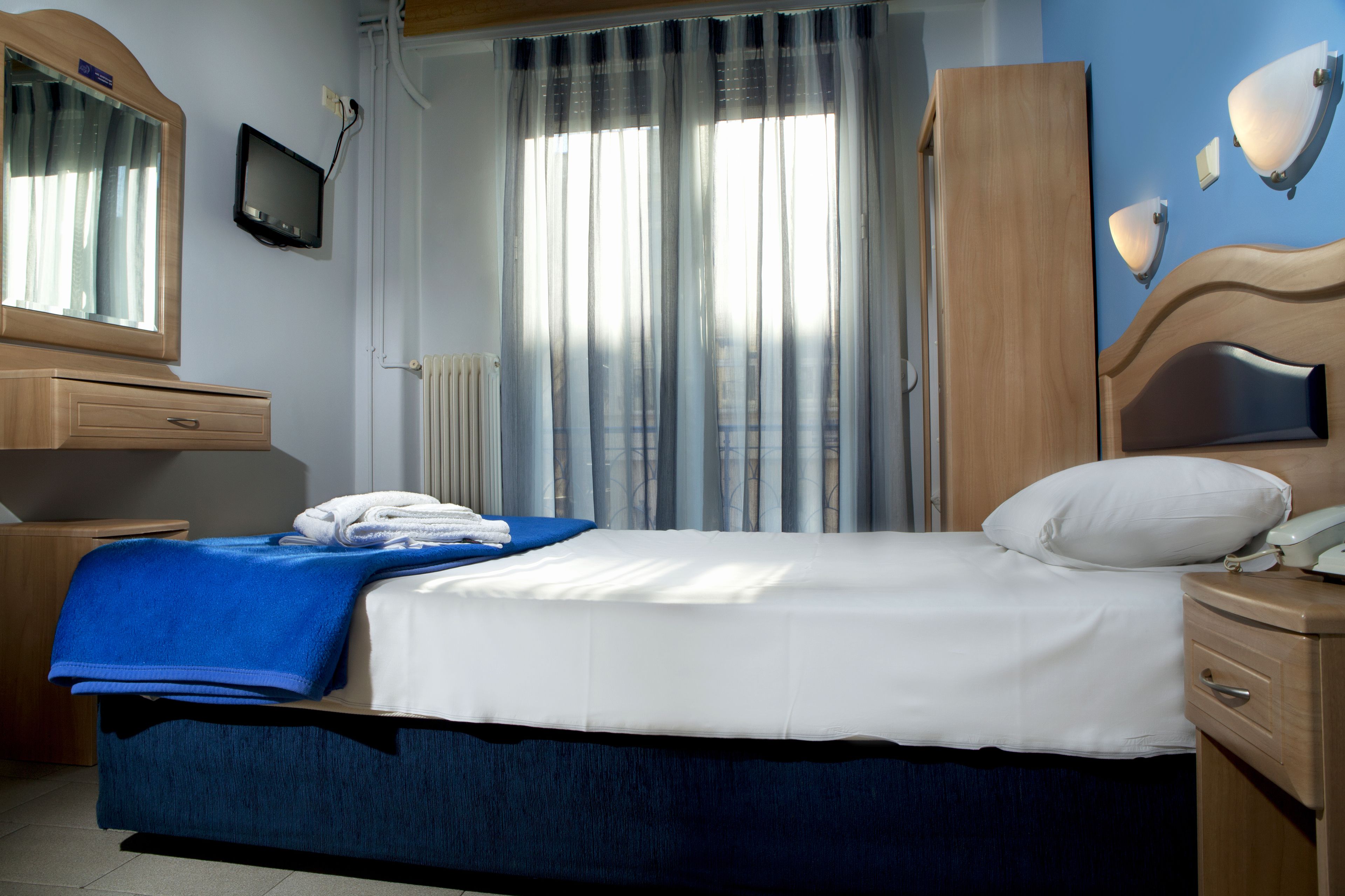 Una cama bien hecha con ropa de cama blanca y azul, una mesita de noche de madera con una lámpara, un armario de madera y un televisor en la pared.
