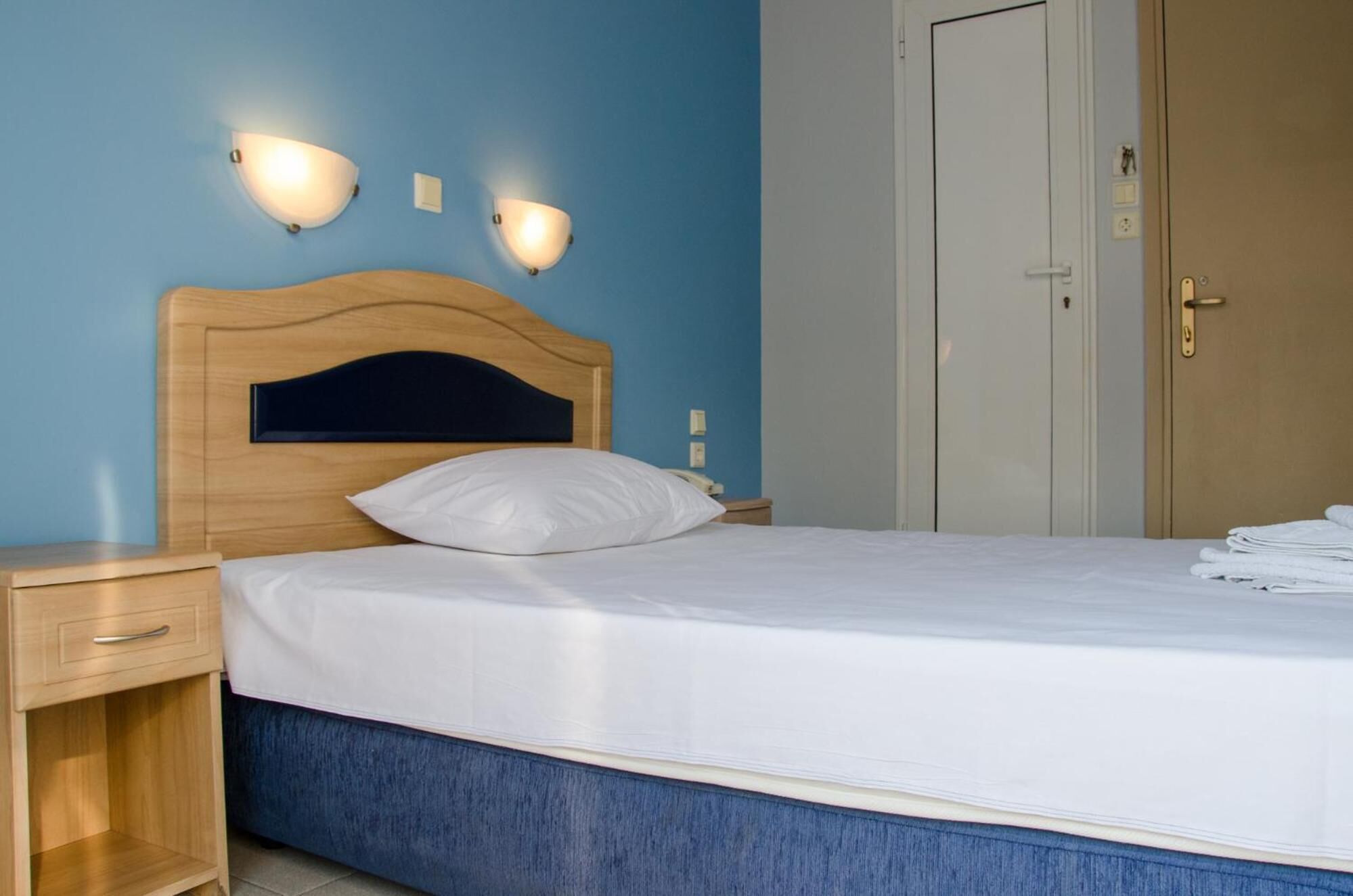 A neatly made bed with white linens, a wooden headboard, and two wall-mounted light fixtures.