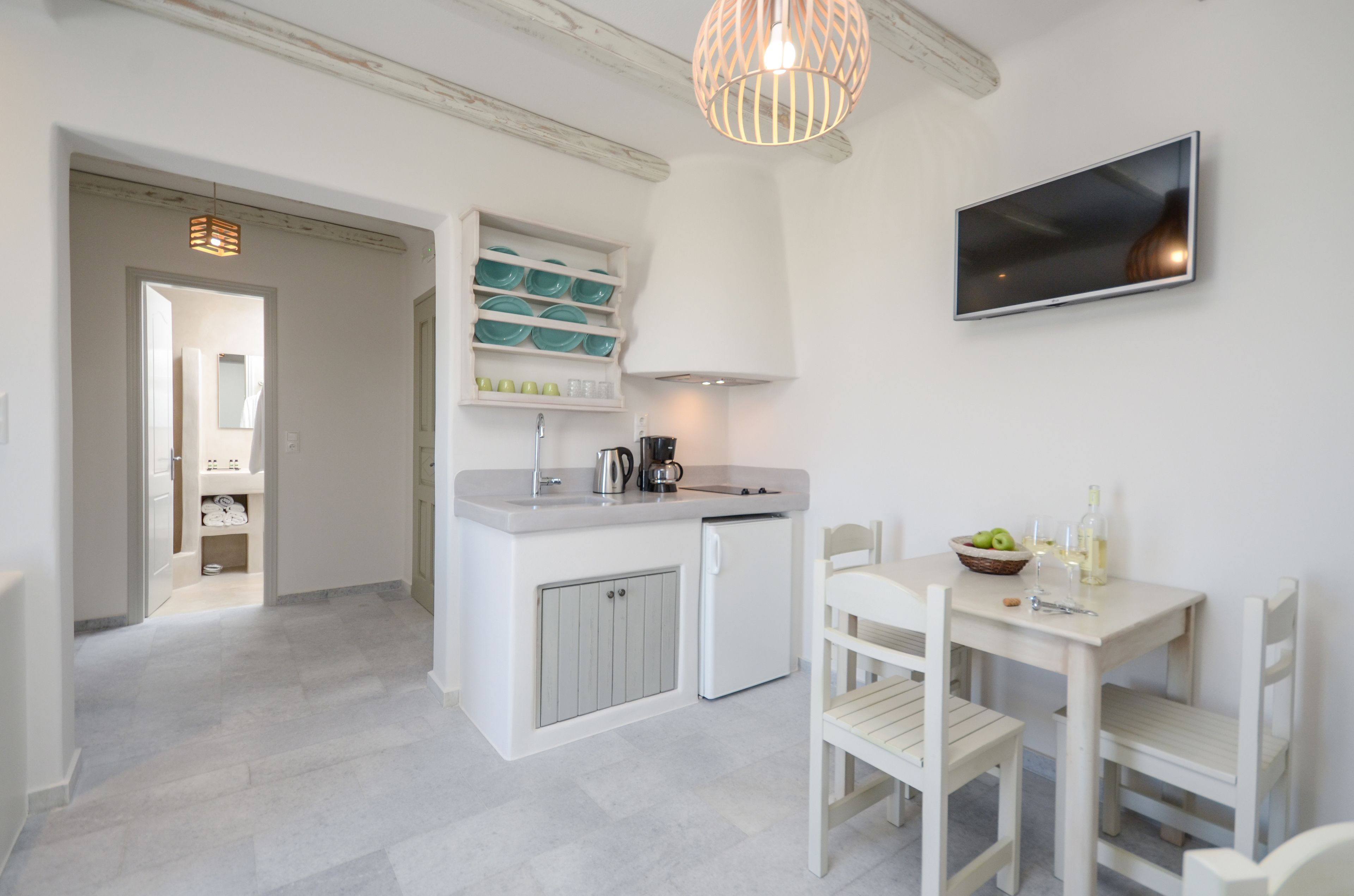 Une cuisine compacte avec un plan de travail blanc, un évier encastré et un petit réfrigérateur. On y trouve une table à manger avec des chaises blanches et un saladier rempli de fruits. Un téléviseur est fixé au mur.