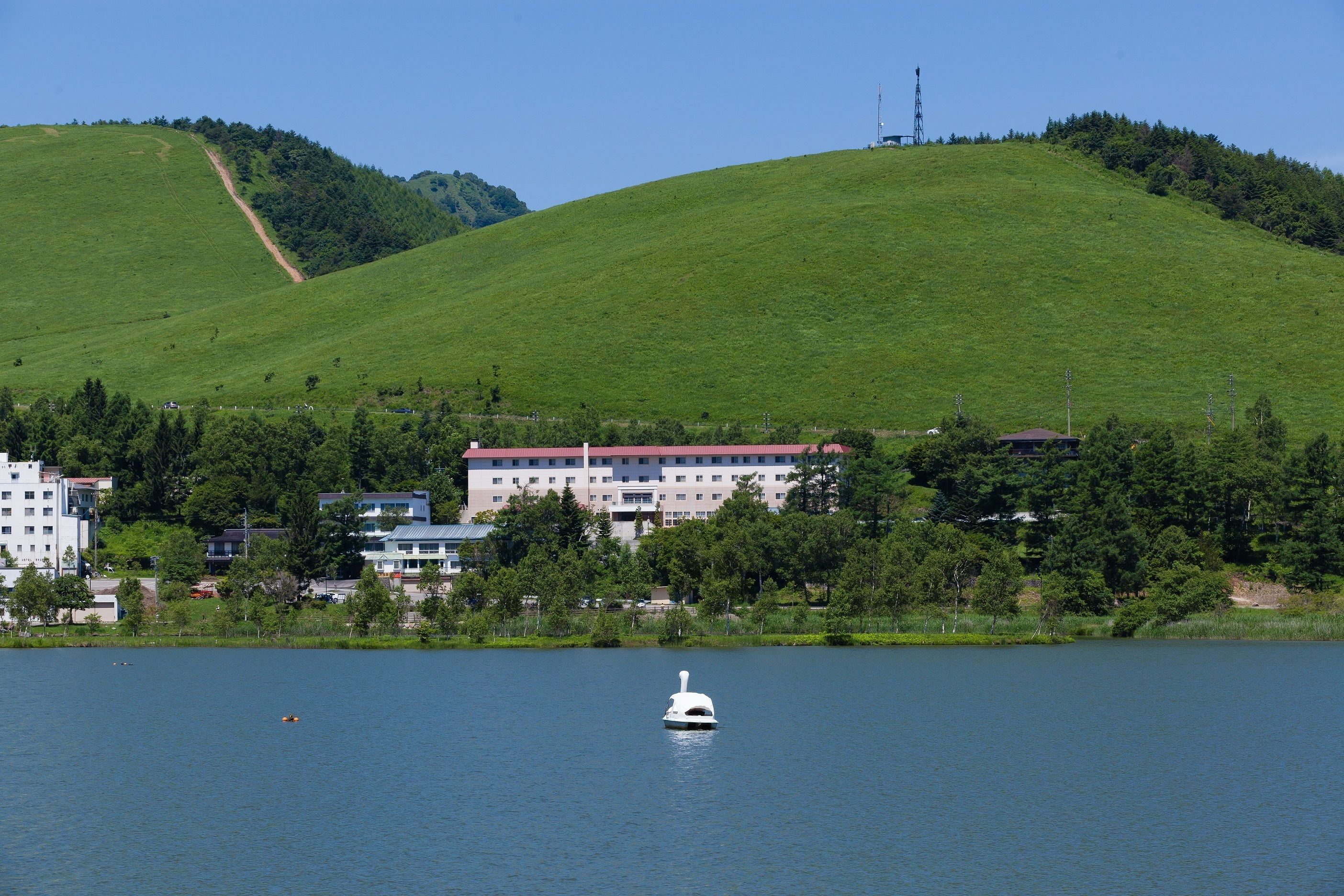 A lake with a white boat in the foreground, surrounded by a green hill and buildings in the background.