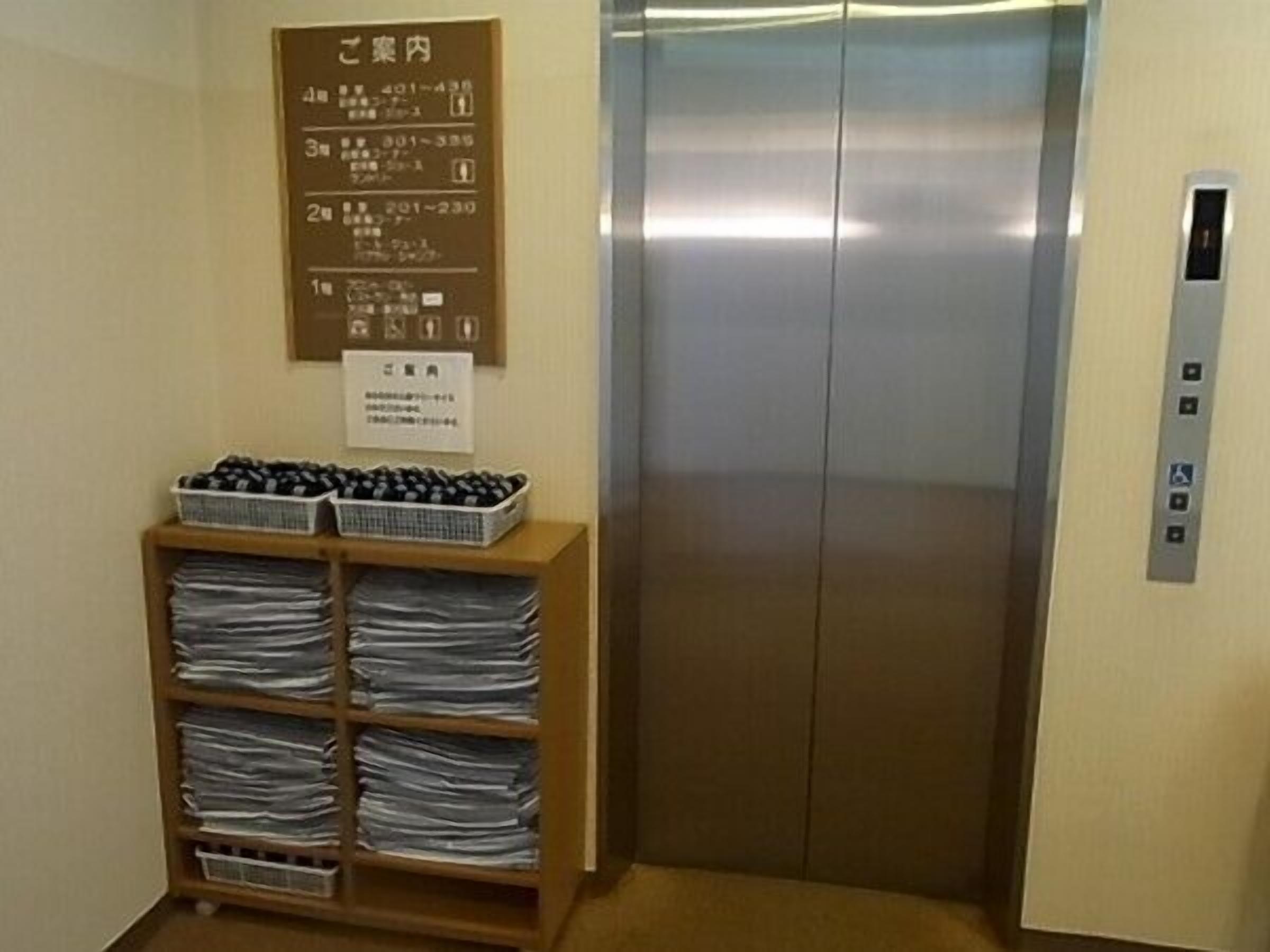 Un ascensor con panel de control, un estante de madera con cestas y papeles, y un cartel en la pared con texto japonés.