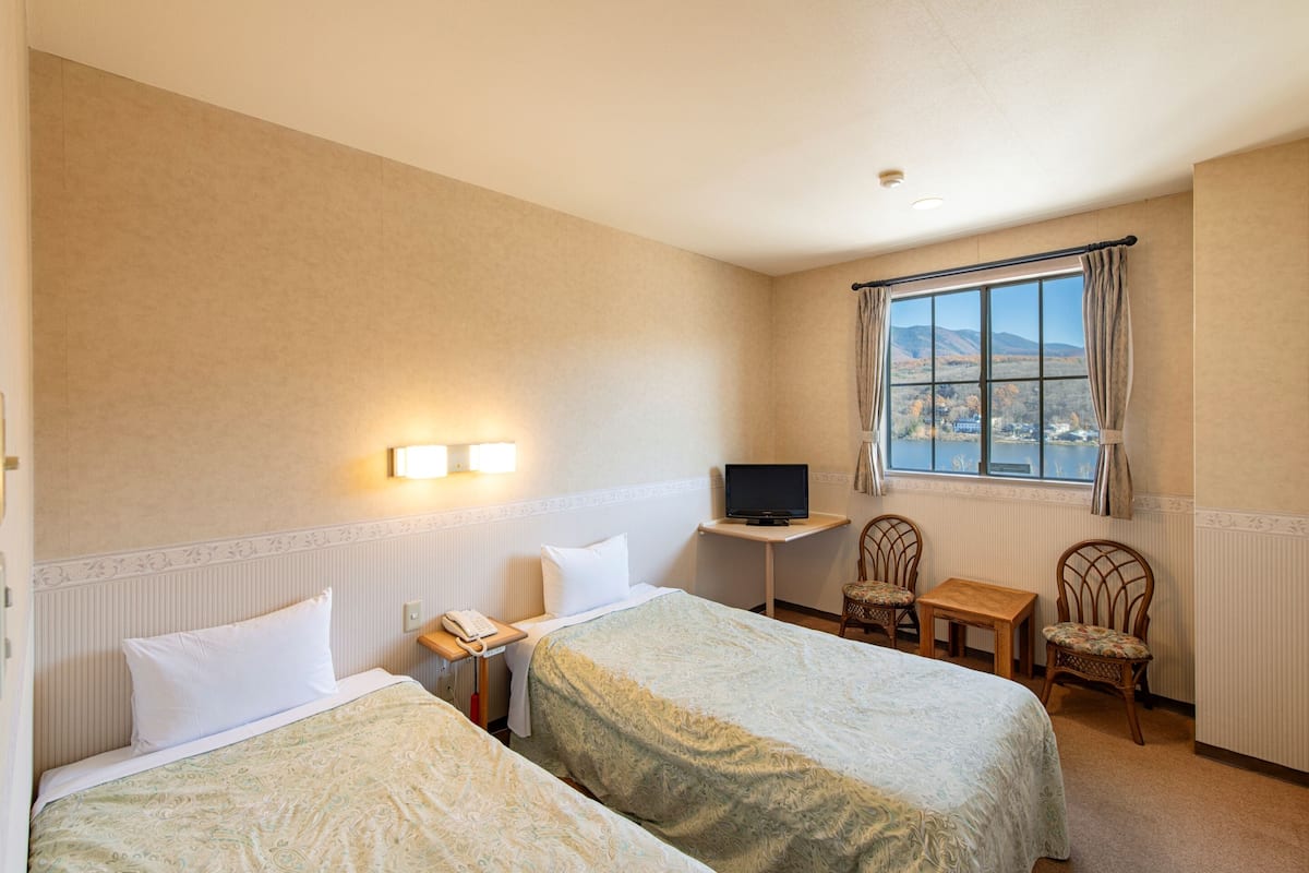Ein Hotelzimmer mit zwei Betten, einem Fernseher, einem kleinen Tisch, zwei Stühlen und einem Fenster mit Blick auf einen Berg und einen See.