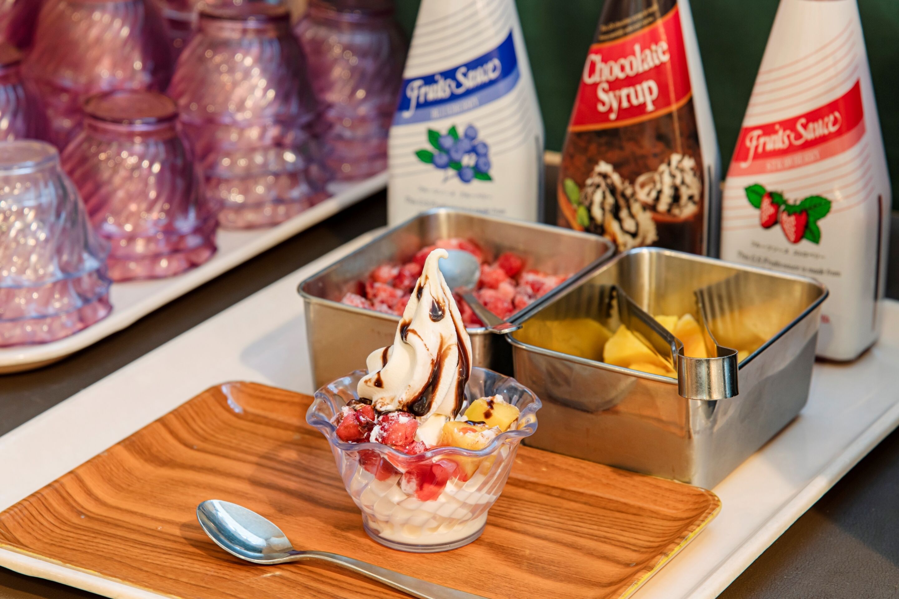 A dessert station with a bowl of ice cream topped with fruits and chocolate syrup, accompanied by bottles of fruit sauce and chocolate syrup.
