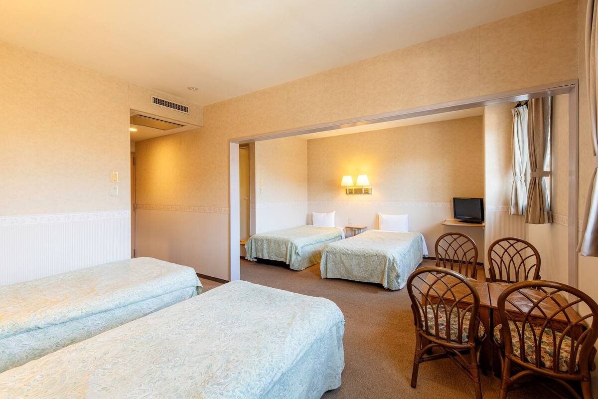 Ein Hotelzimmer mit zwei Einzelbetten, einem Esstisch mit vier Stühlen, einem Fernseher und einem sichtbaren Badezimmer hinter einer offenen Tür.