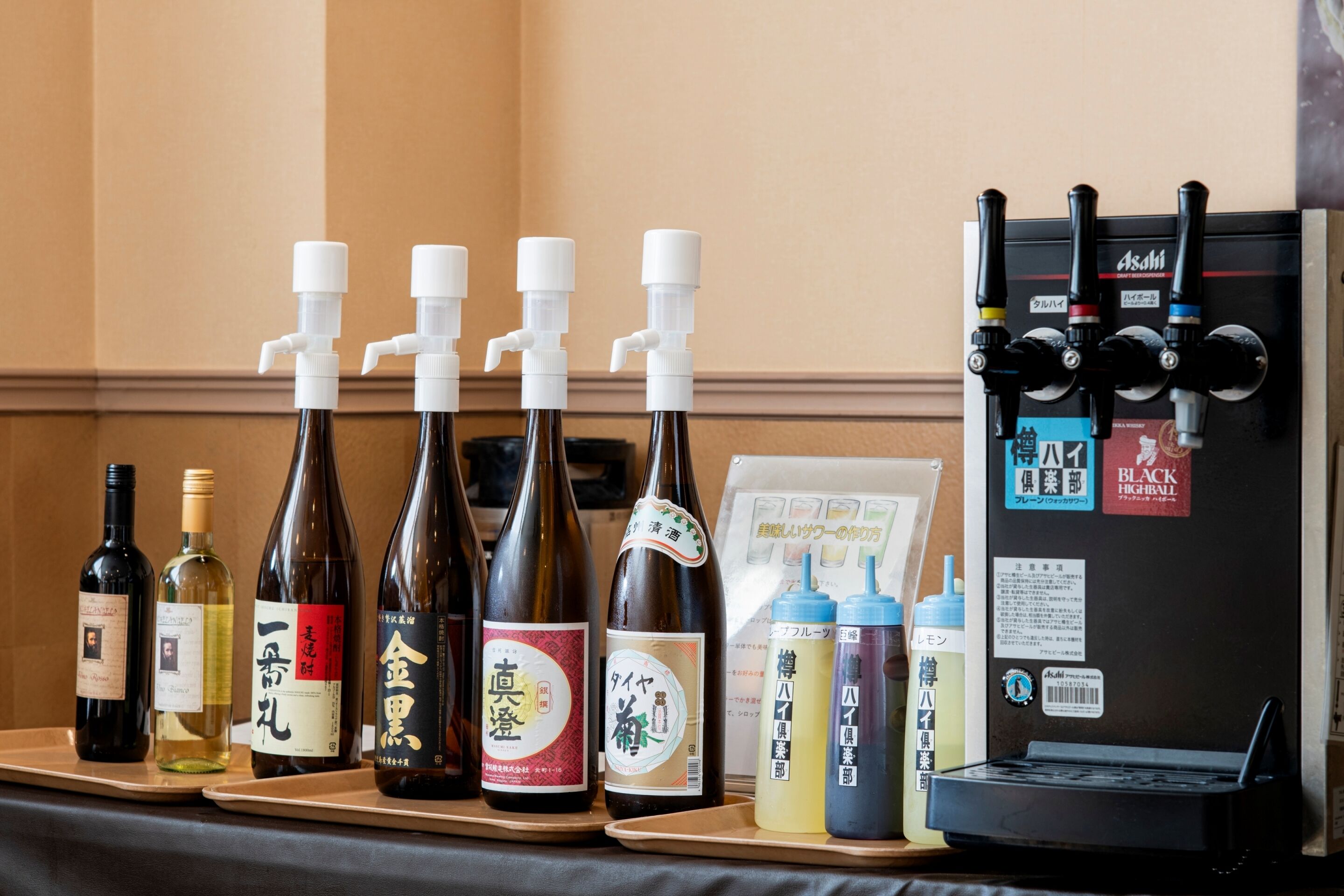 A selection of bottles on a tray, including various types of alcoholic beverages and condiments, with a refrigerator featuring multiple handles and a menu on the wall.
