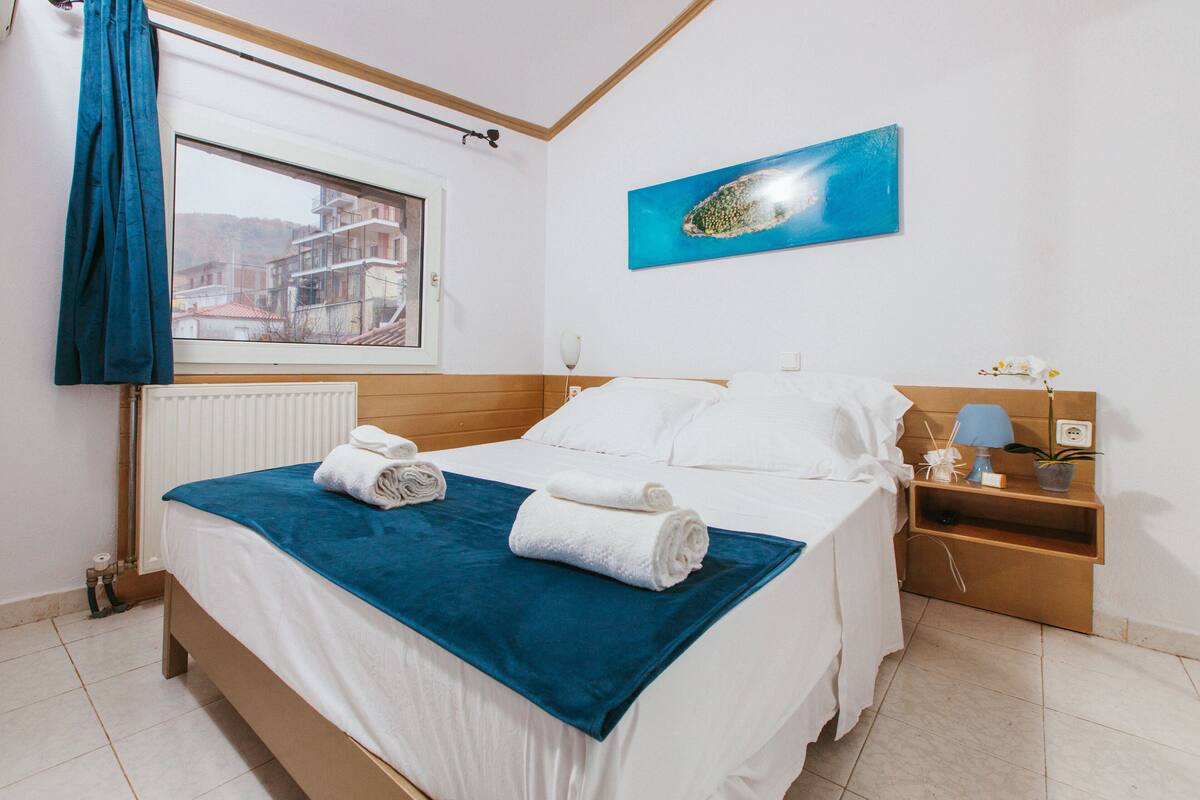 Une chambre d’hôtel avec un lit, une fenêtre donnant sur des immeubles, une œuvre d’art bleue accrochée au mur, et une table de chevet avec une lampe et une plante.