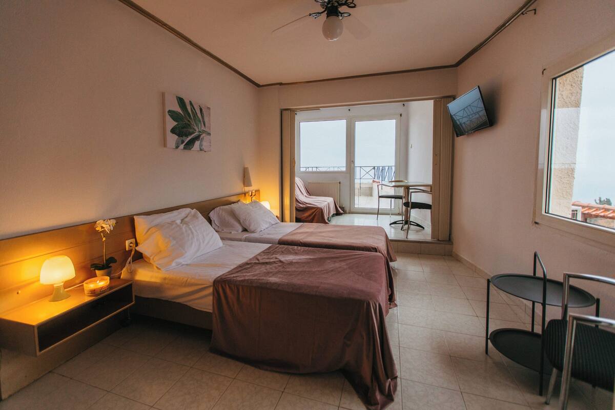Une chambre d’hôtel avec deux lits, une petite table, une chaise, un téléviseur fixé au mur et un balcon équipé d’une chaise et d’une table.