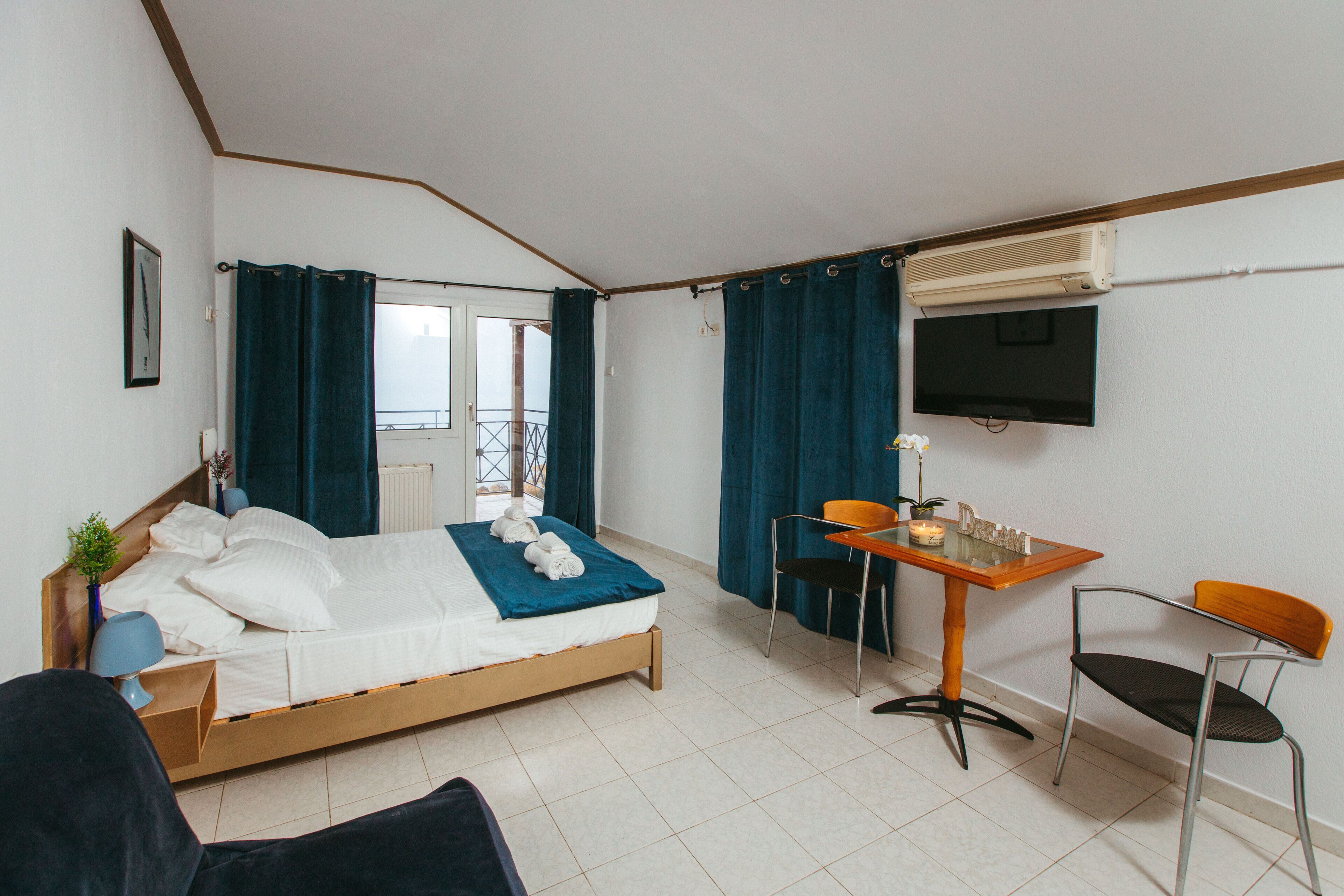 A bedroom with a bed, a desk, and a chair. There is a window with curtains, a TV mounted on the wall, and a plant on the bedside table.