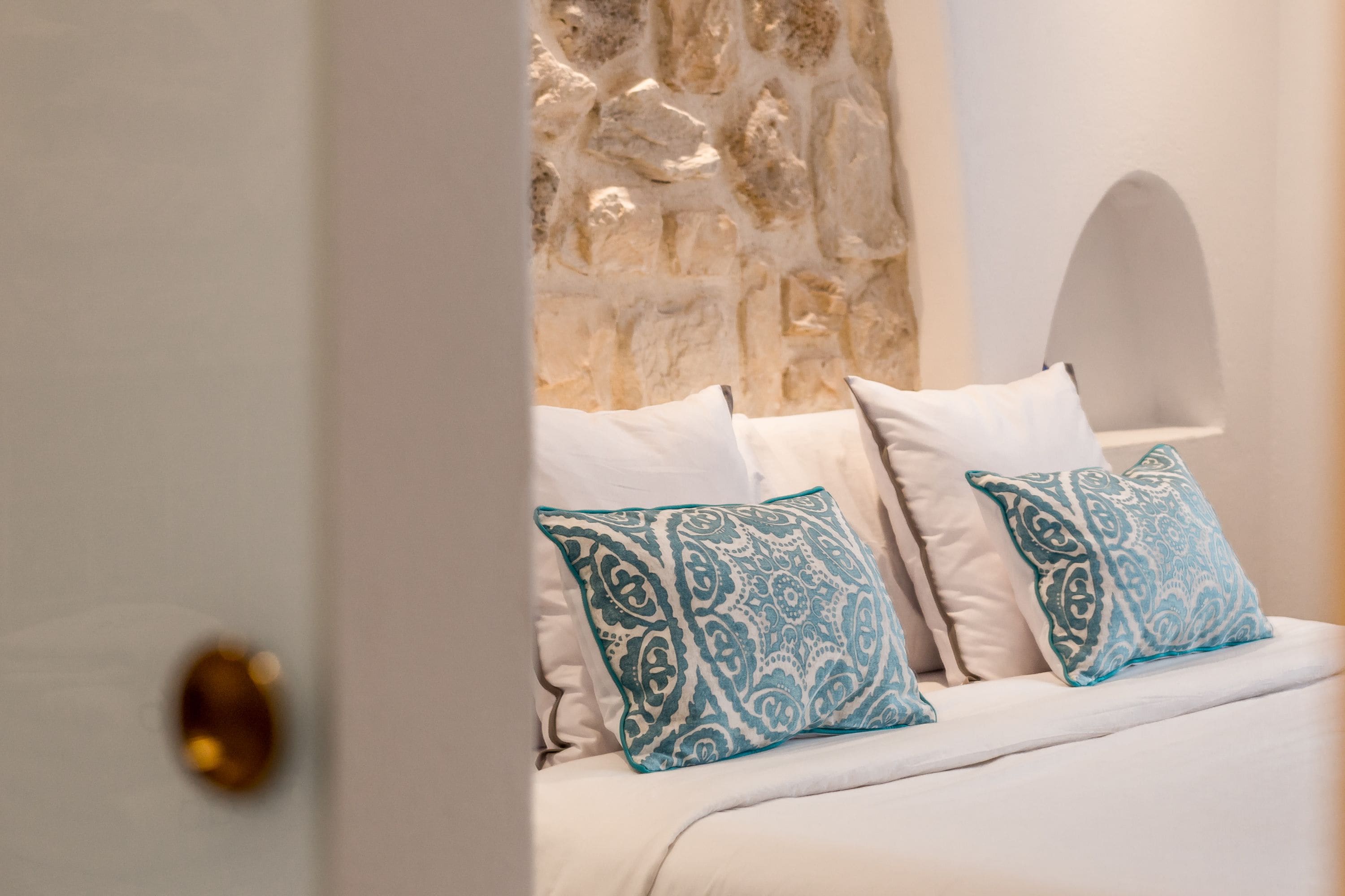 A bed with white bedding and blue patterned pillows, a stone wall, and a white arched window.