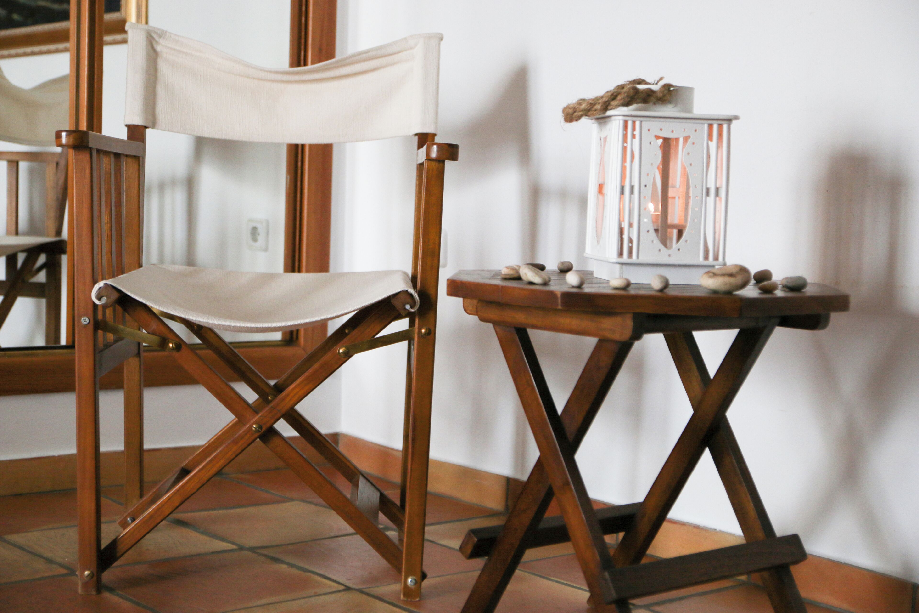 Uma cadeira de madeira com assento e encosto de lona, uma pequena mesa de madeira com um candeeiro e um chão de azulejos.