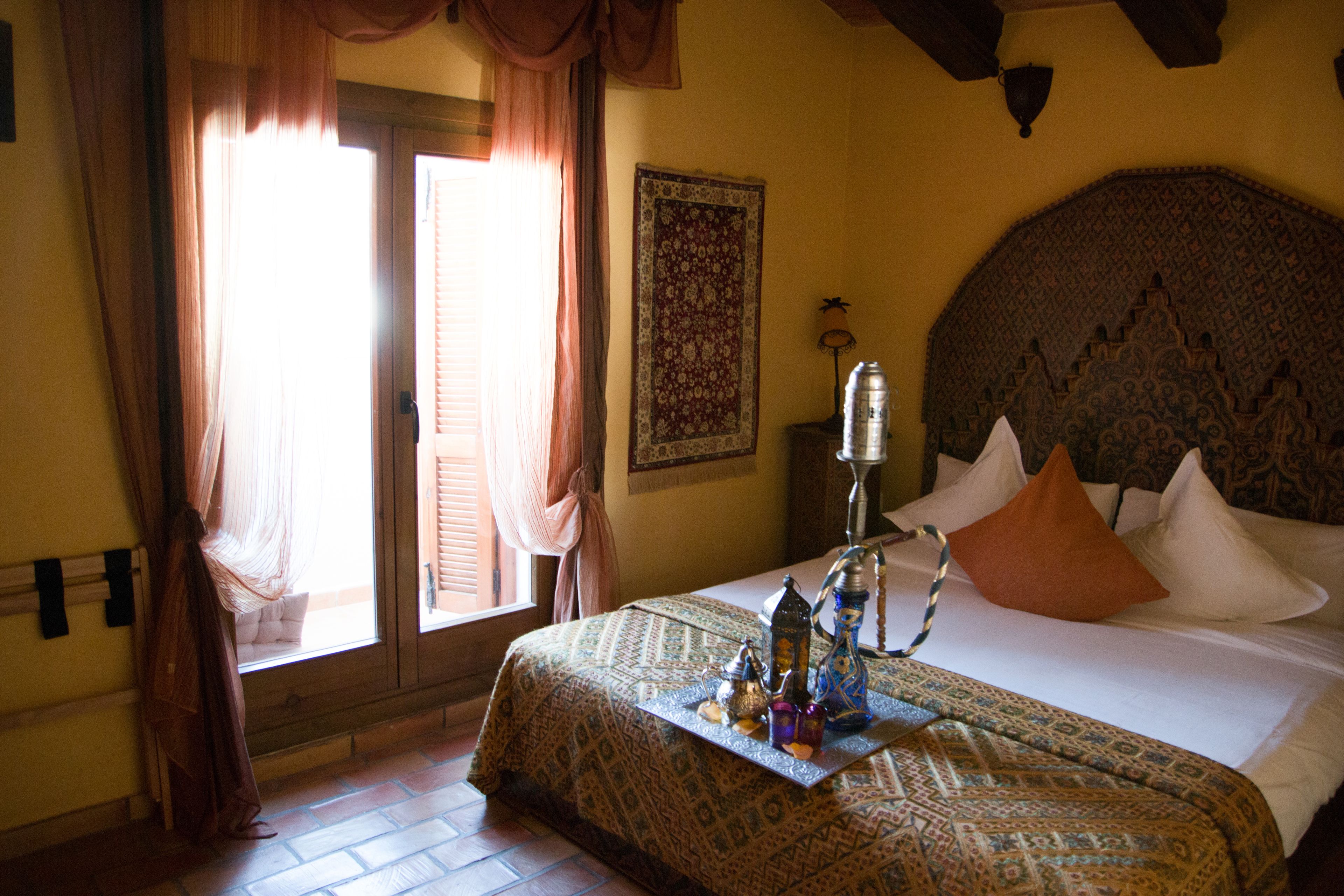 A hotel room with a bed, a window with curtains, a wall-mounted lamp, and a decorative headboard.