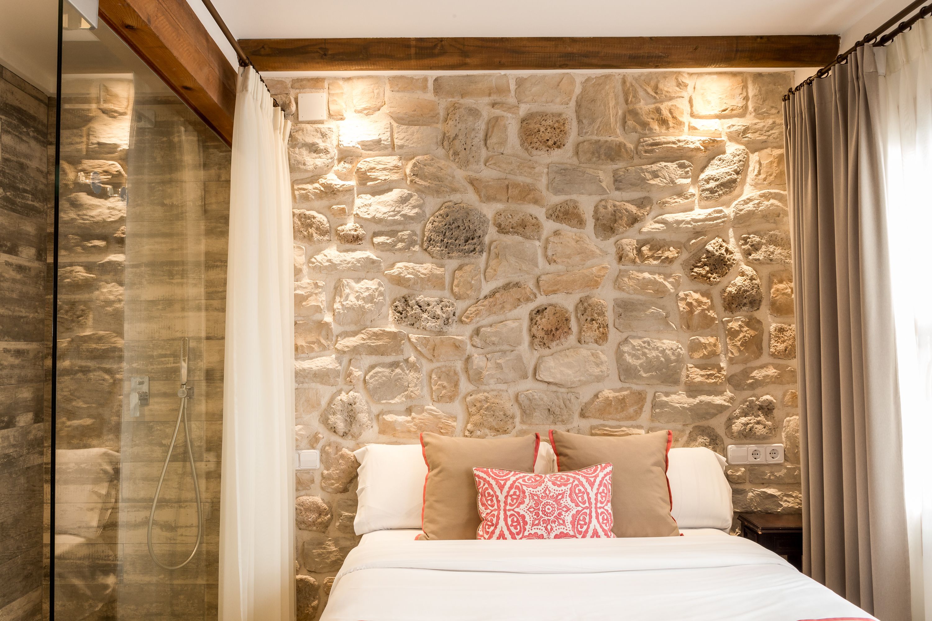 A bedroom with a stone wall, a bed with white and red bedding, and a shower area.