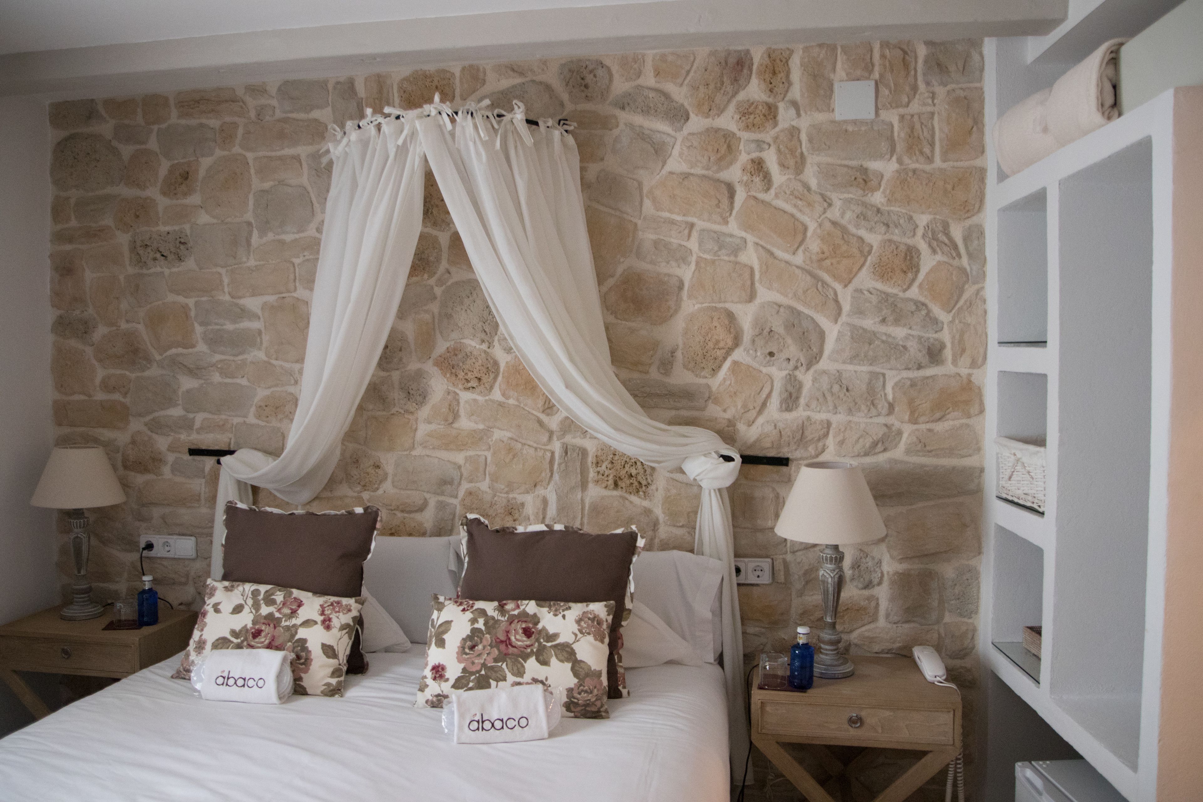 Une chambre avec un mur en pierre, un lit à baldaquin, deux tables de chevet avec des lampes et une petite table de nuit avec un téléphone et des bouteilles.