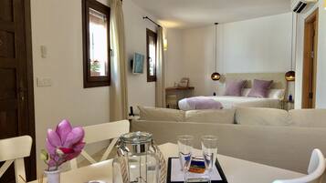 A hotel room with a bed, a sofa, a dining table with two glasses and a menu, a vase with flowers, and a wall-mounted air conditioner.