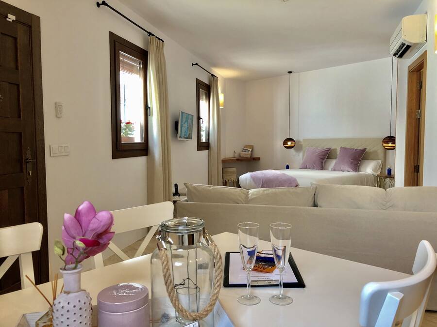 A hotel room with a bed, a sofa, a dining table with two glasses and a menu, a vase with flowers, and a wall-mounted air conditioner.