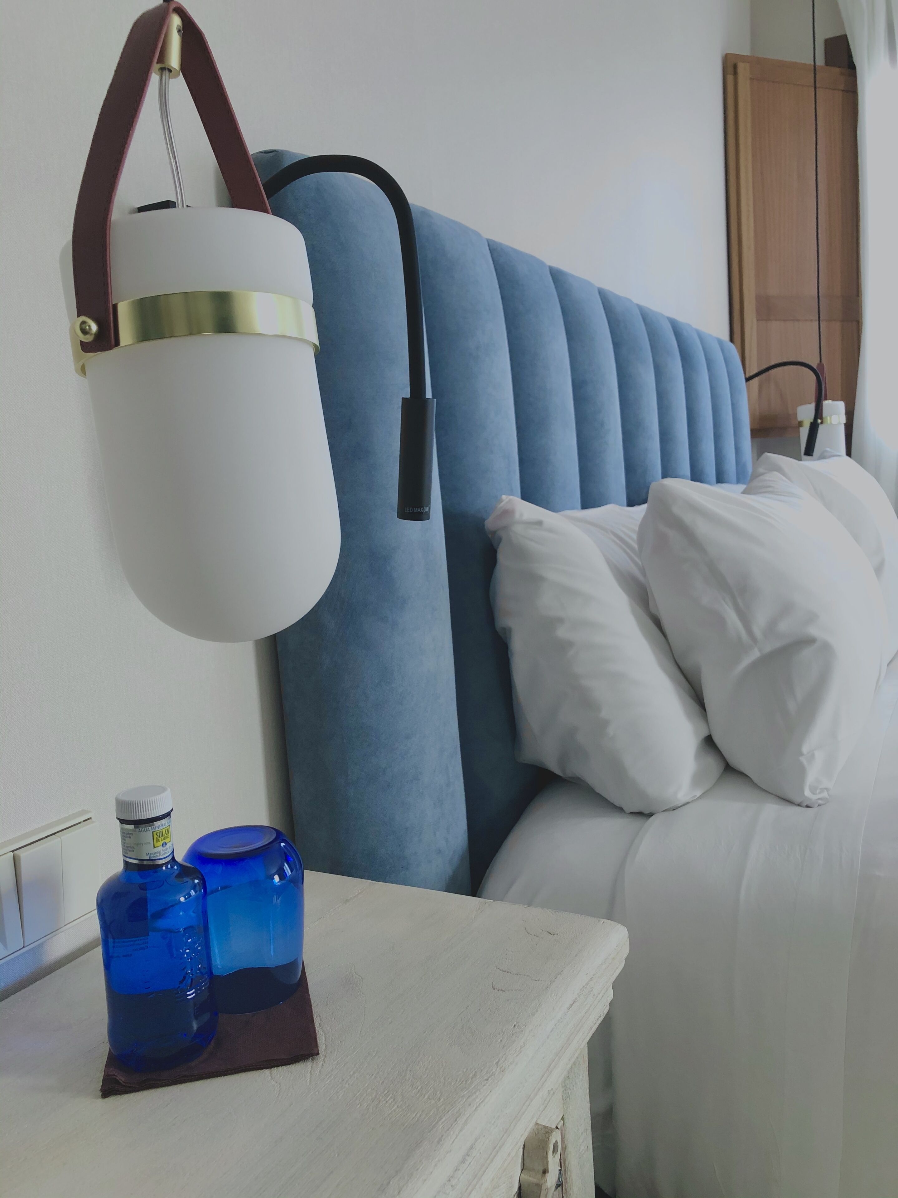 Un dormitorio con cabecera capitonada, una mesita de noche con lámpara y un mueble auxiliar con dos botellas.