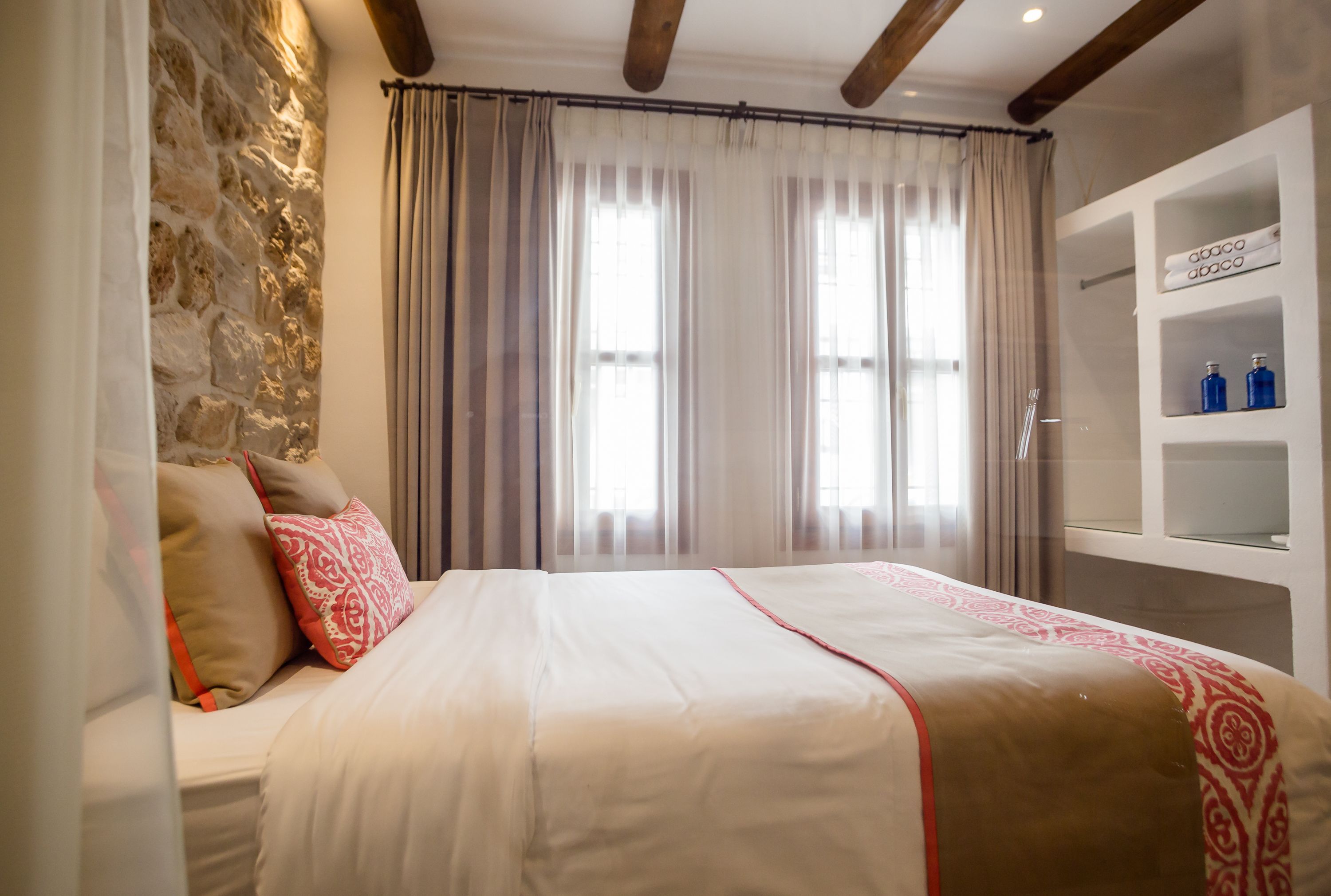 A bedroom with a stone wall, a bed with patterned bedding, a window with curtains, and a shelf with toiletries.