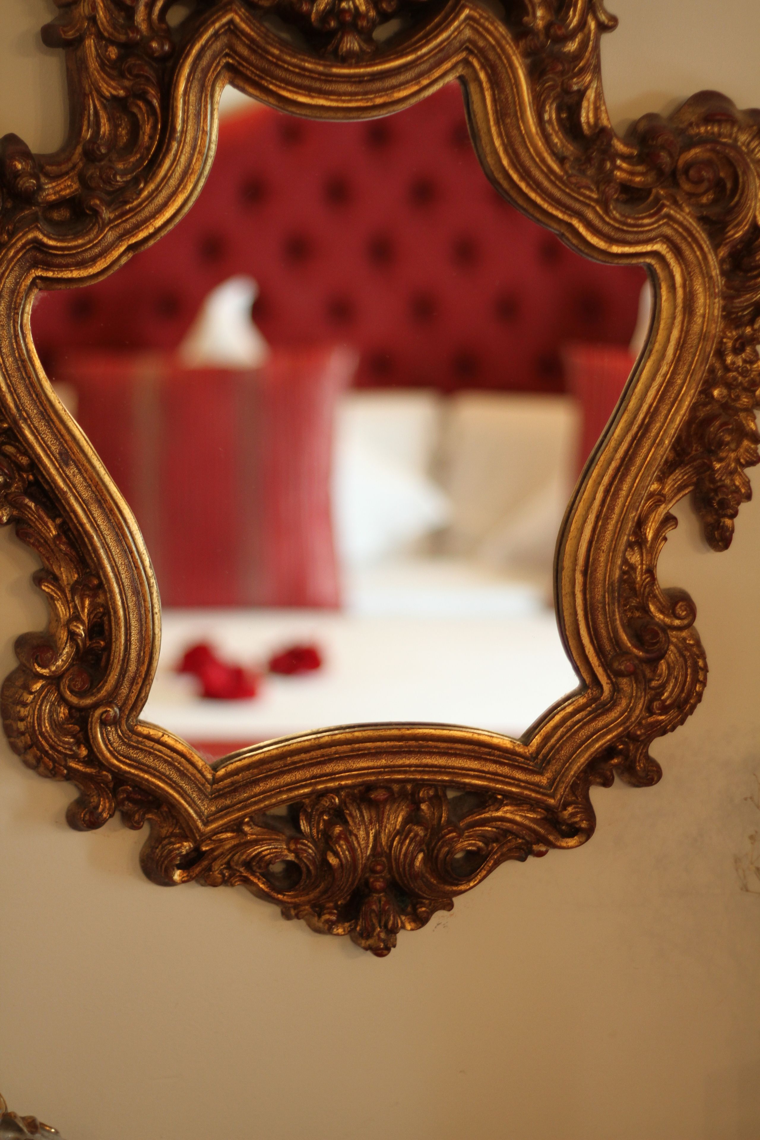 A mirror with an ornate gold frame reflecting a red upholstered sofa and a white bed with red pillows.