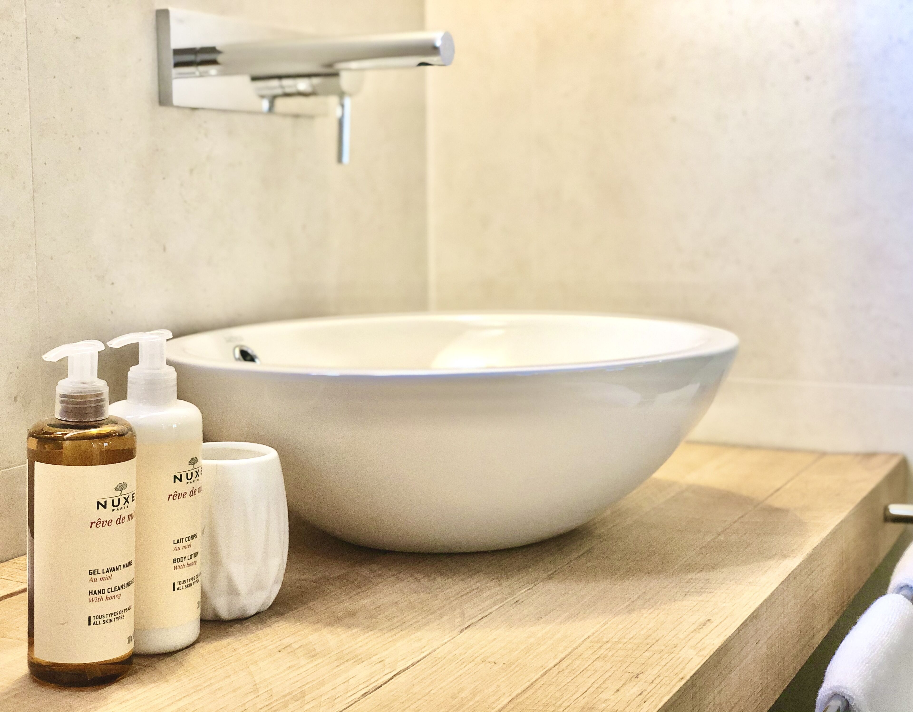 A bathroom with a white sink, wooden countertop, and two bottles of Nuxe body lotion.