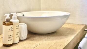 A bathroom with a white sink, wooden countertop, and two bottles of Nuxe body lotion.