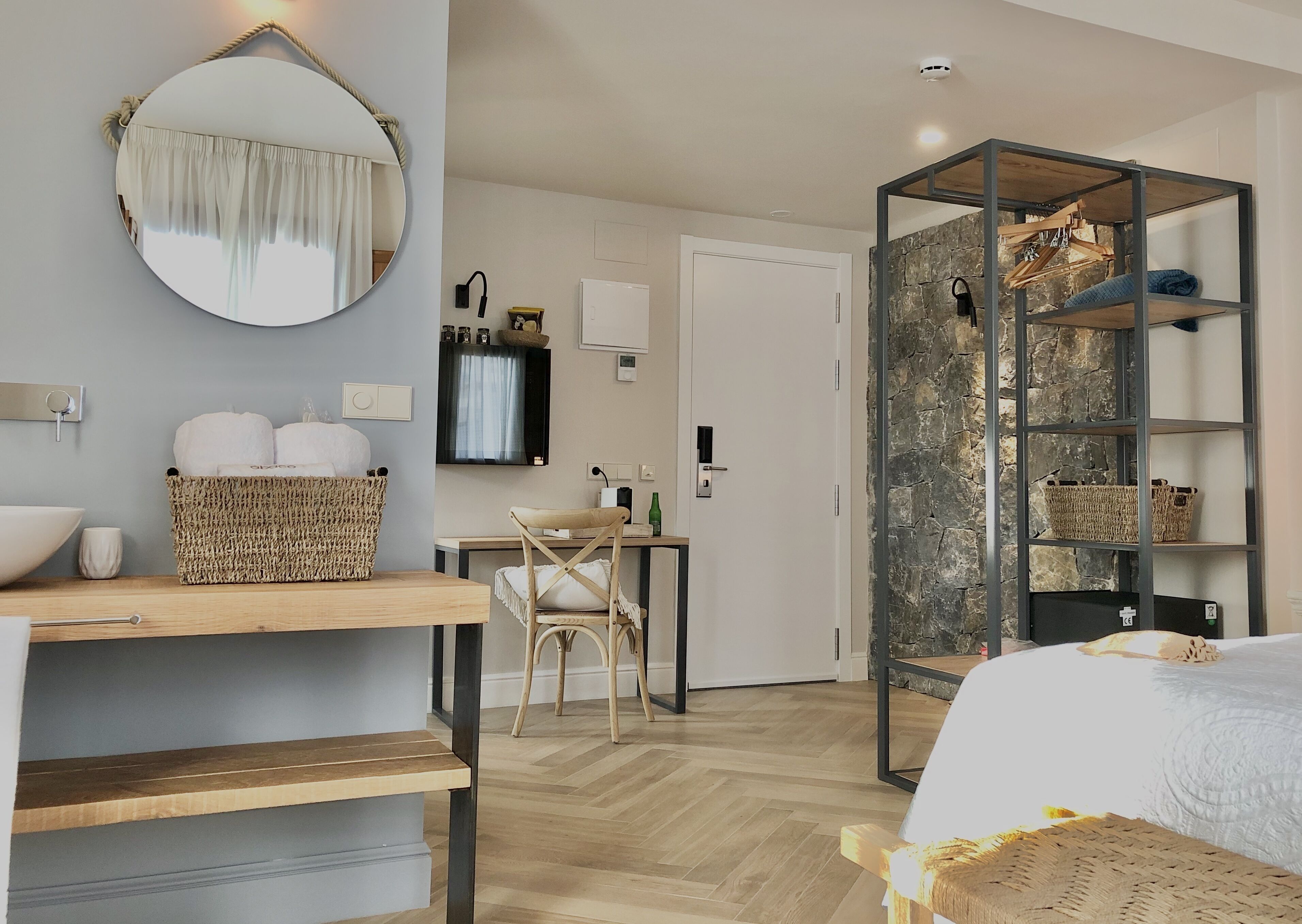 Un hotel moderno con un escritorio de madera, un espejo redondo, una cama con cabecera blanca, una pared de acento de piedra y una estantería empotrada.