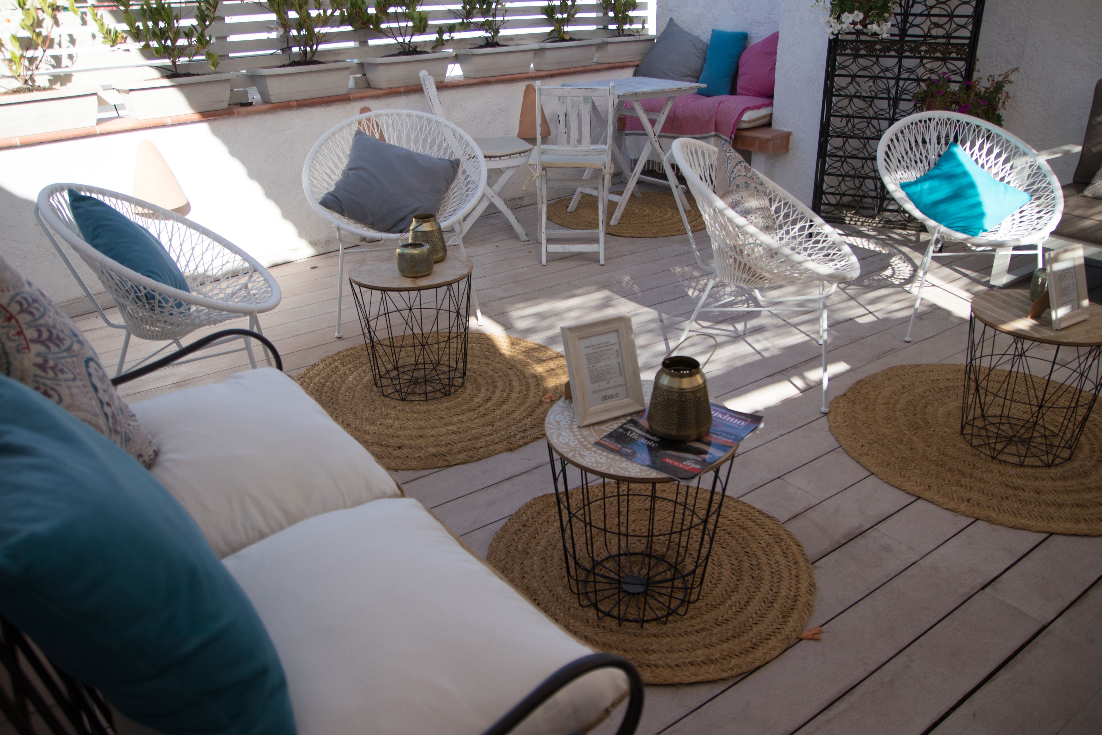 Eine sonnendurchflutete Terrasse mit weißen Korbstühlen, gewebten Matten und einem kleinen Tisch mit einer Zeitschrift.