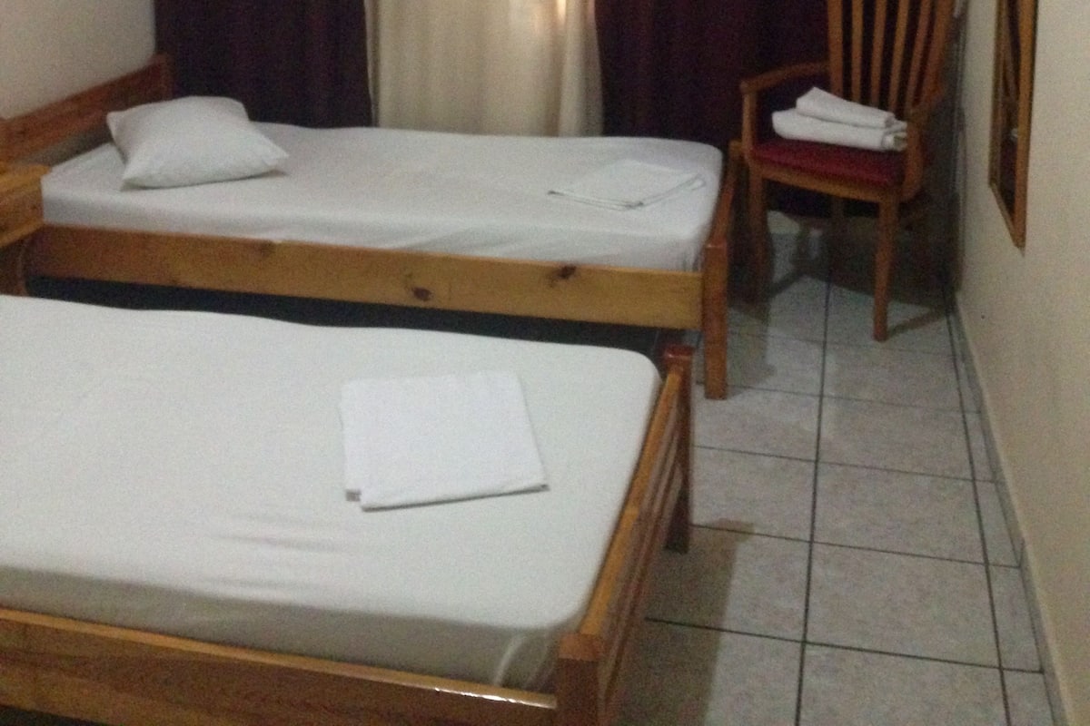 A room with two single beds, a chair, a window with curtains, and a wall-mounted light fixture.