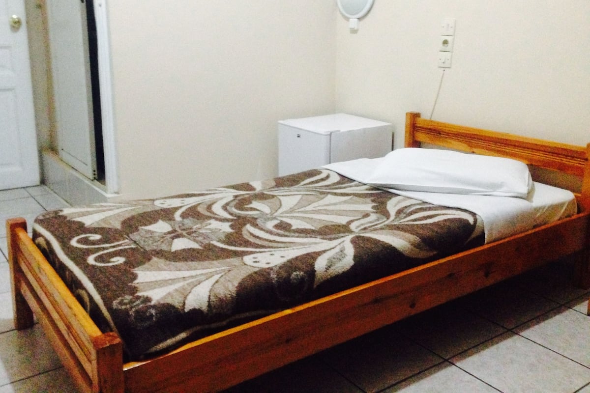 A single bed with a patterned bedspread, a wooden headboard, a white bedside table, and a wall-mounted light fixture.