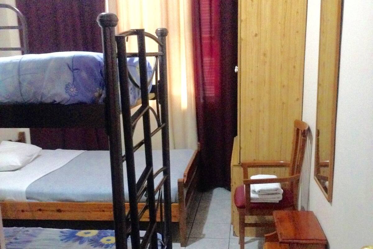 A small, compact room with a bunk bed, a wooden chair, a small wooden table, and a mirror on the wall.