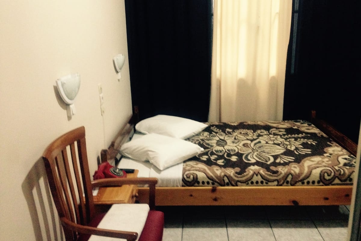 A single bed with a patterned bedspread, a wooden chair, a small table, and a wall-mounted light fixture.
