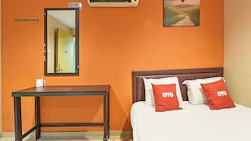 A hotel room with an orange wall, a bed with two pillows, a bedside table with a tissue box, a mirror, and an air conditioning unit.