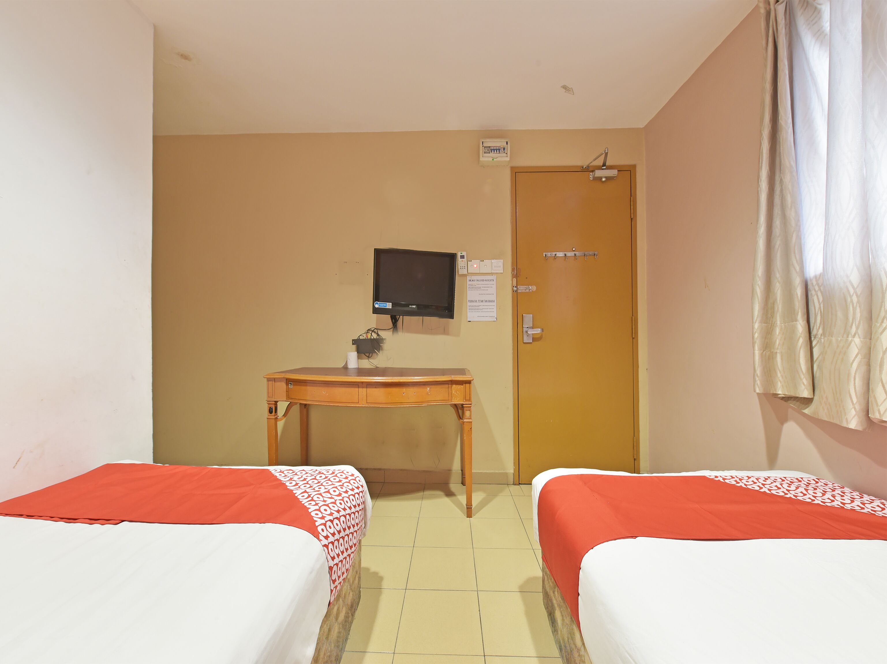 A hotel room with two single beds, a small wooden desk, a television mounted on the wall, and a door with a security lock.