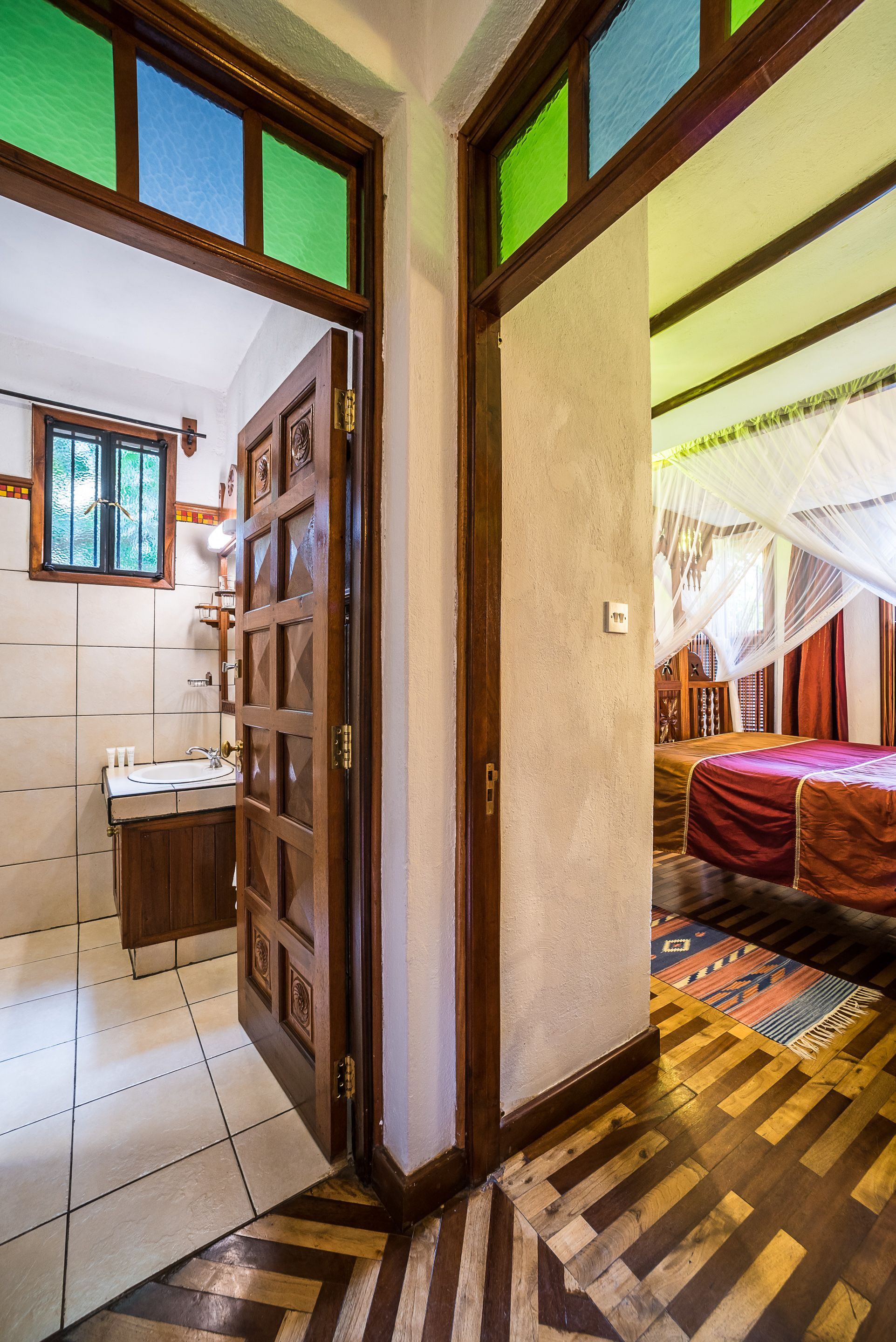Una habitación con una puerta de madera que da a un baño con lavamanos e inodoro. La habitación tiene una cama con colcha roja y una alfombra estampada en el suelo.