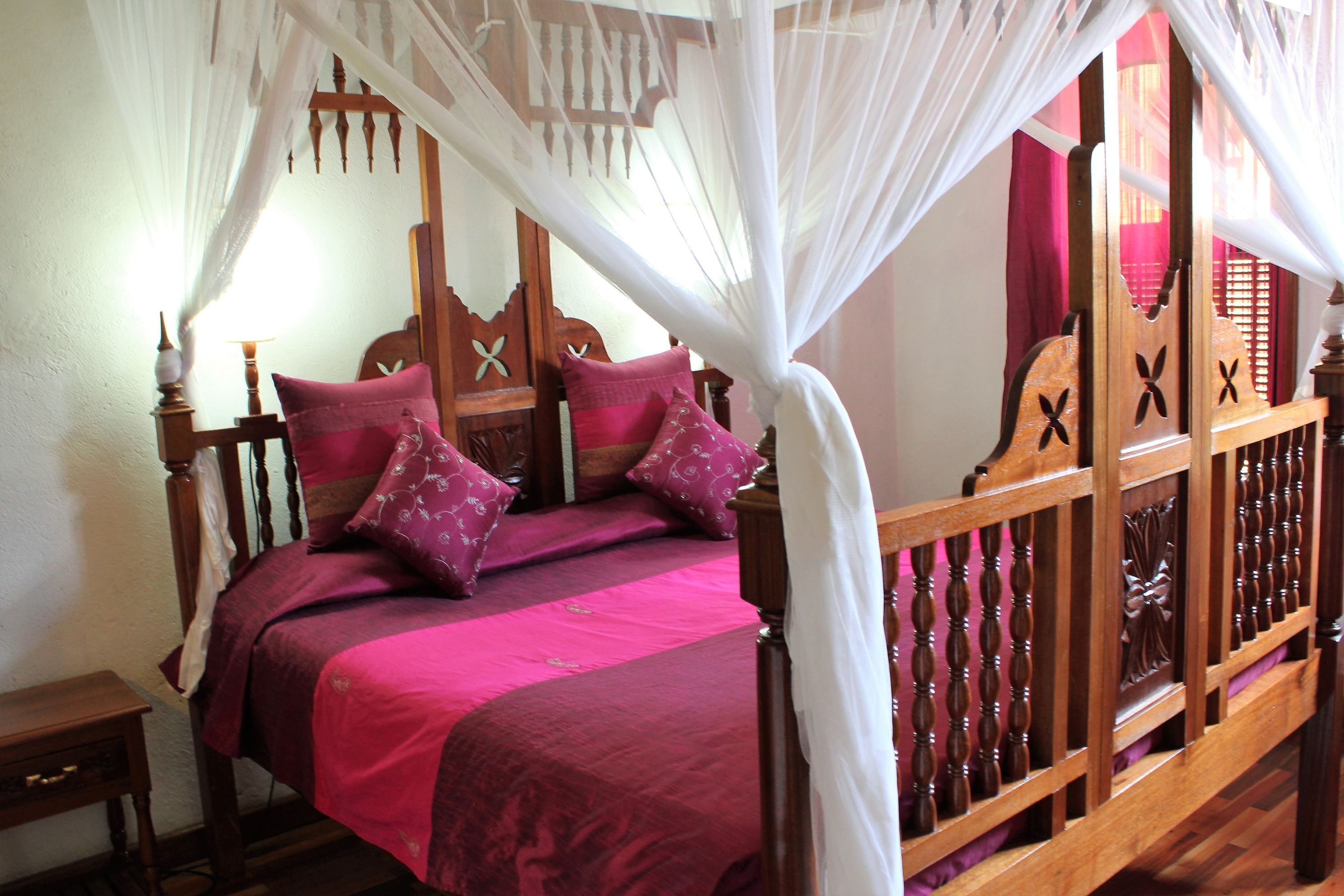 A bed with a canopy, wooden headboard with X-shaped carvings, and a pink bedspread.