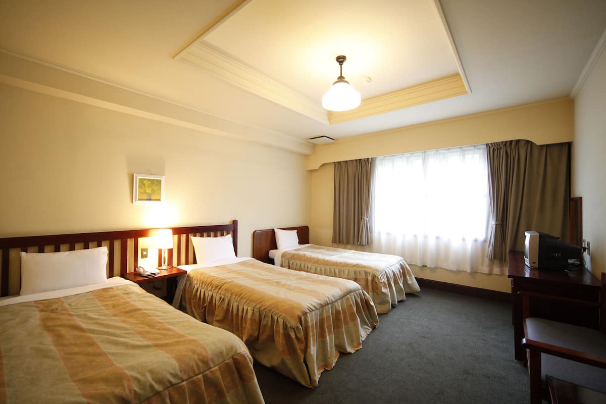 Quarto de hotel com duas camas de solteiro, uma escrivaninha com cadeira, ventoinha no teto e janela com cortinas.