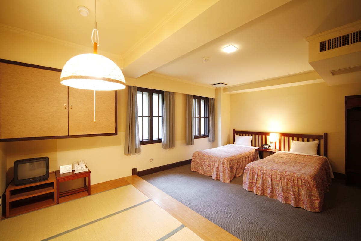 Quarto de hotel com duas camas, televisão, mesa pequena e piso tradicional de tatame japonês.