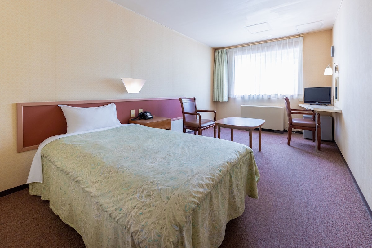 Quarto de hotel com uma cama, uma escrivaninha, uma cadeira e uma televisão.