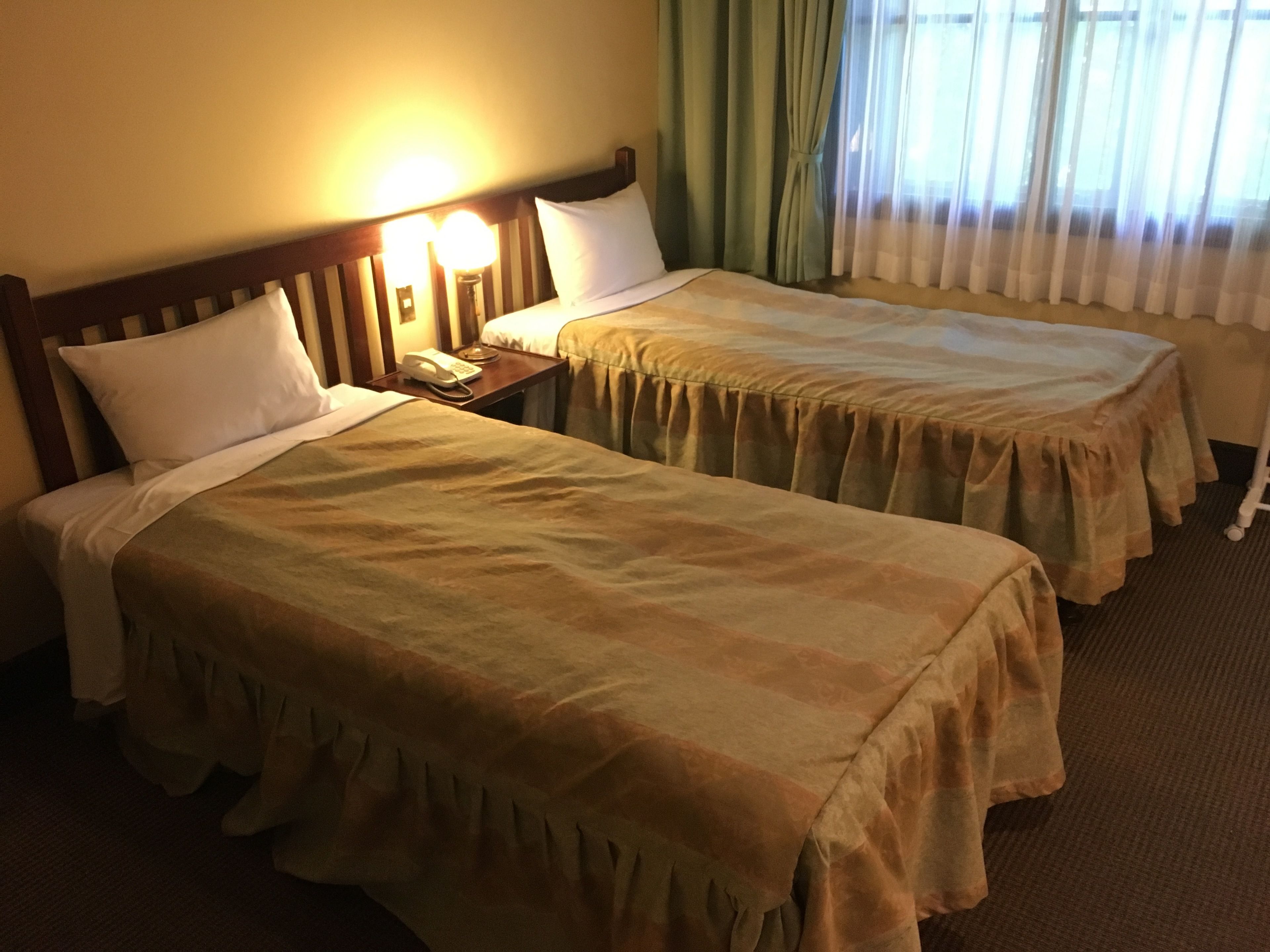A hotel room with two single beds, a bedside table with a lamp, and a window with curtains.