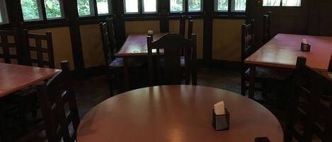 En restaurantindretning med runde borde og stole, store vinduer med udsigt til træer, og et lys på et bord.