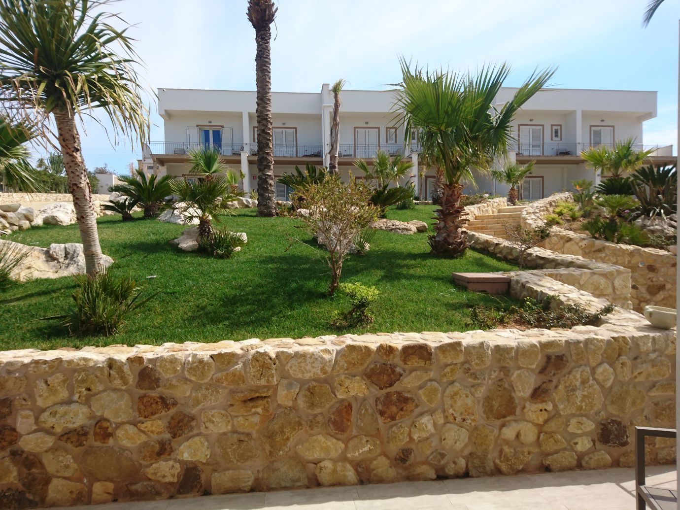 A courtyard with a stone wall, palm trees, and a building with balconies in the background.