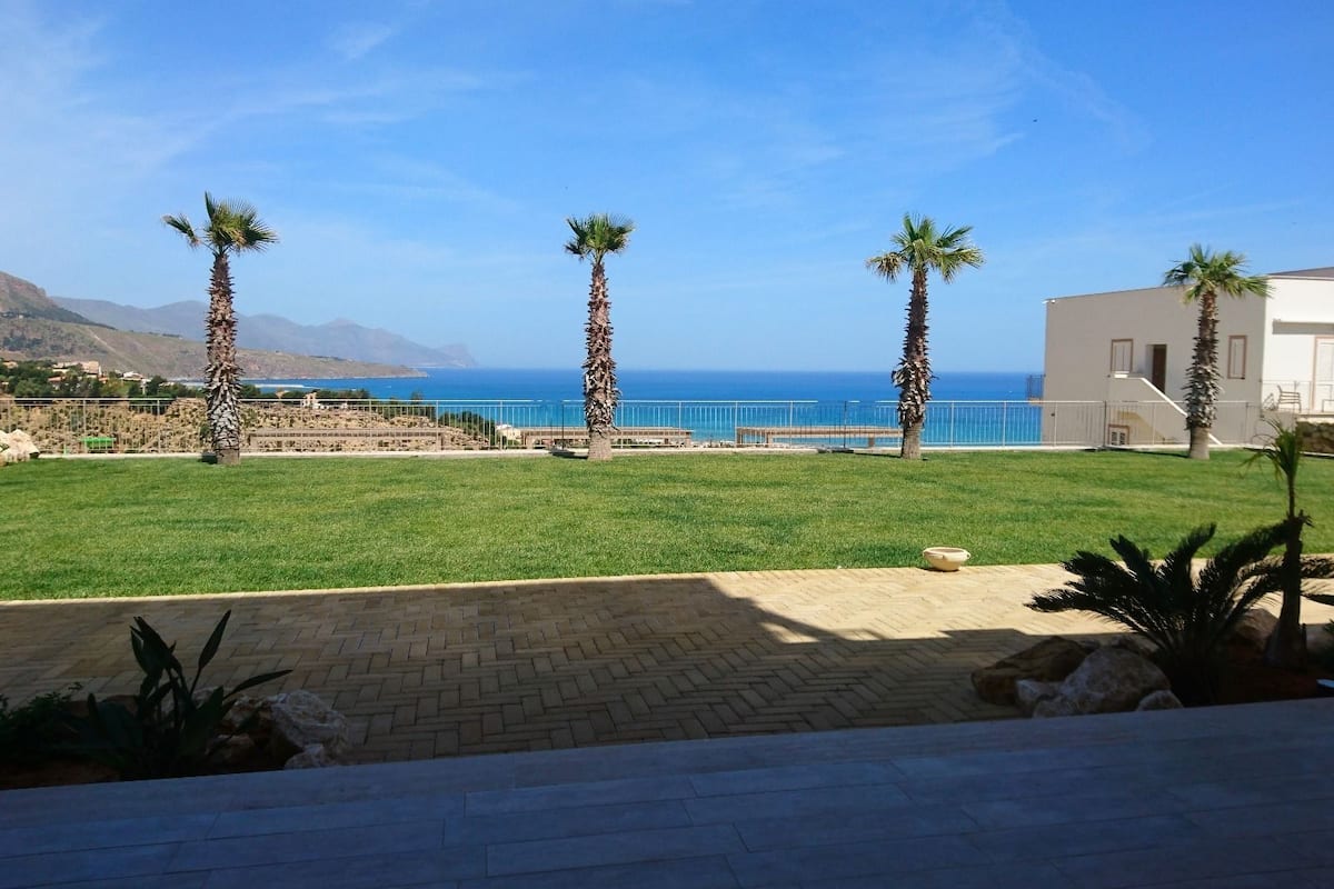 A grassy outdoor area with palm trees, a paved walkway, and a view of the sea.