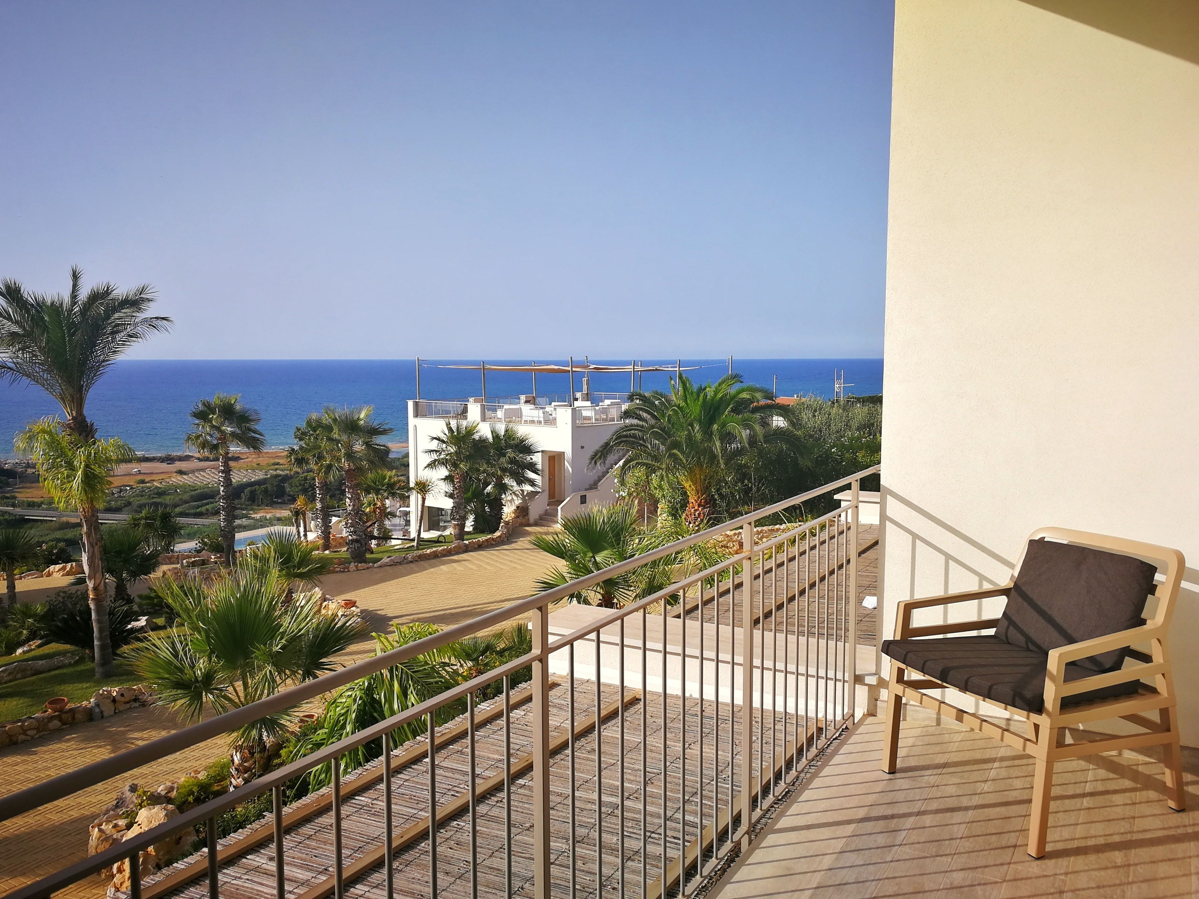 A balcony with a wooden chair, overlooking a beach and a white building with a balcony.