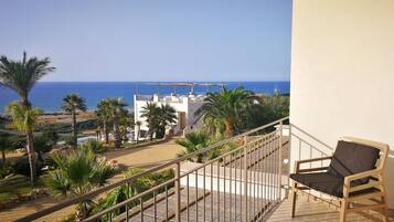 A balcony with a wooden chair, overlooking a beach and a white building with a balcony.