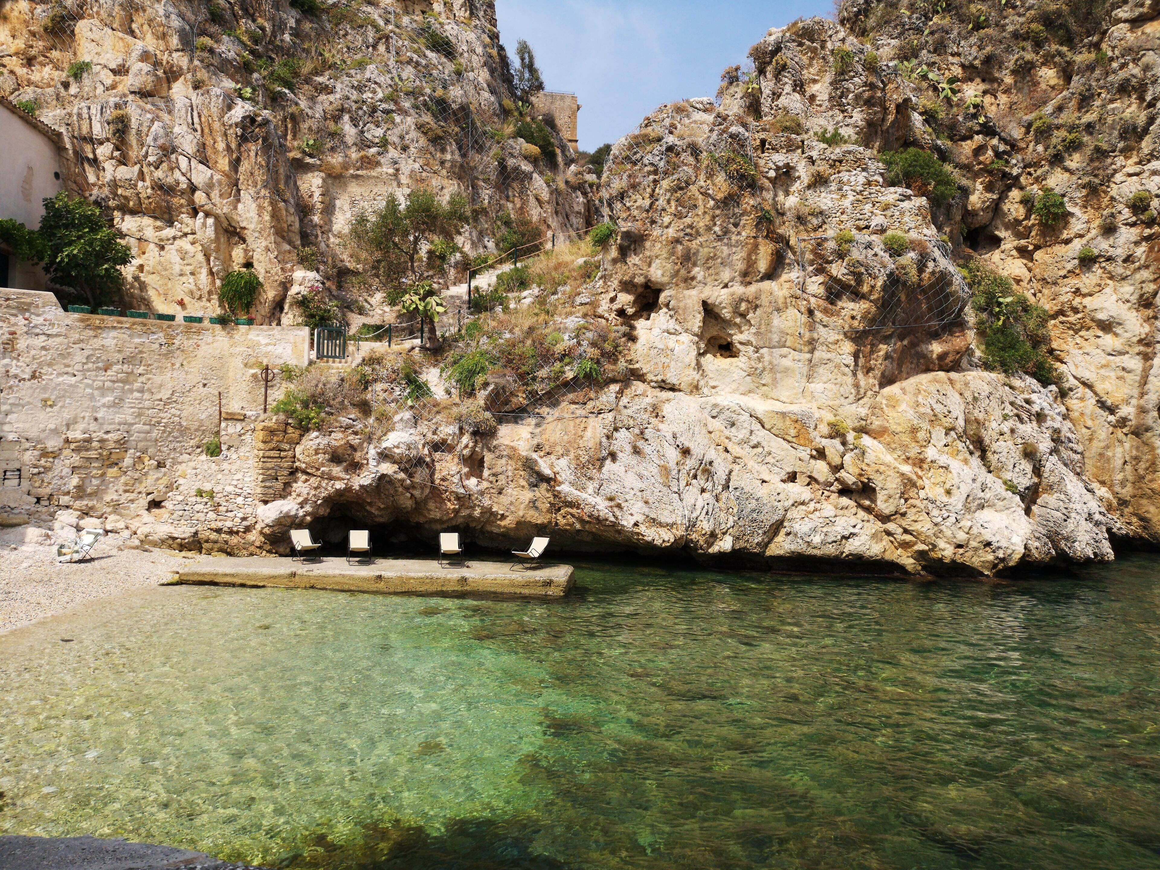 A rocky cliff with clear water, a small dock with chairs, and a building in the background.