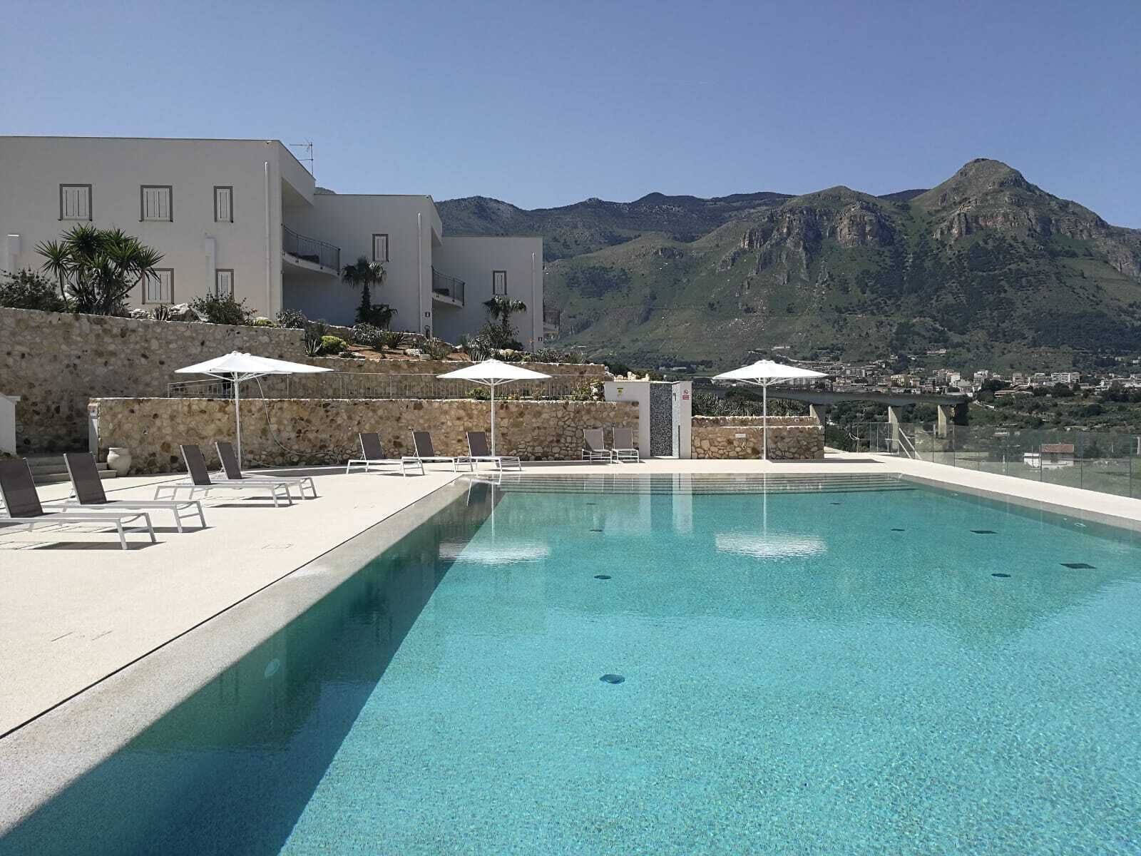 A swimming pool with lounge chairs and umbrellas, surrounded by a stone wall and a building with a mountain view in the background.