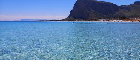 A clear blue sea with a mountain in the background and a beach with buildings.