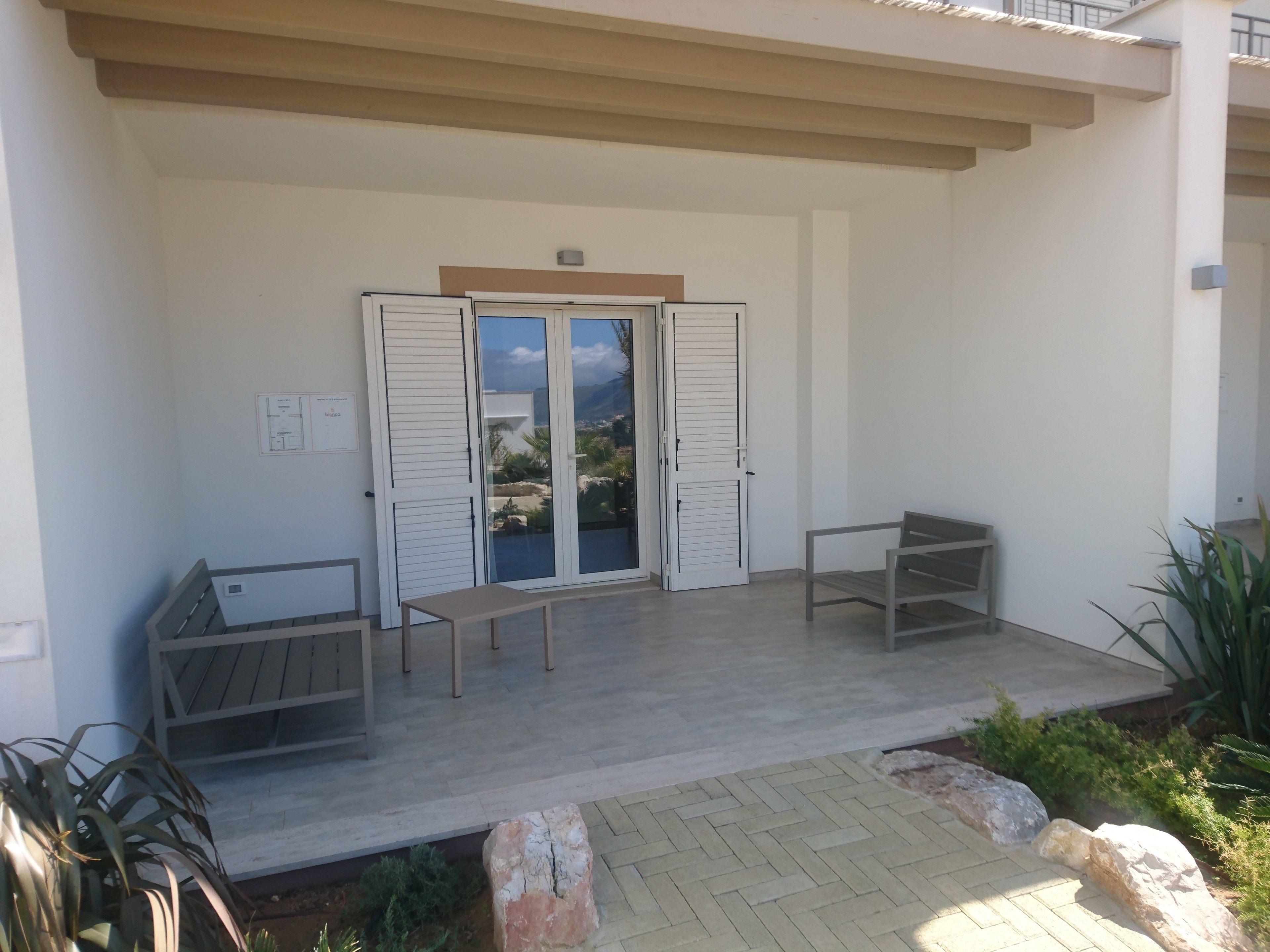 A covered outdoor seating area with two benches and a table, a sliding glass door, and a view of the surrounding landscape.